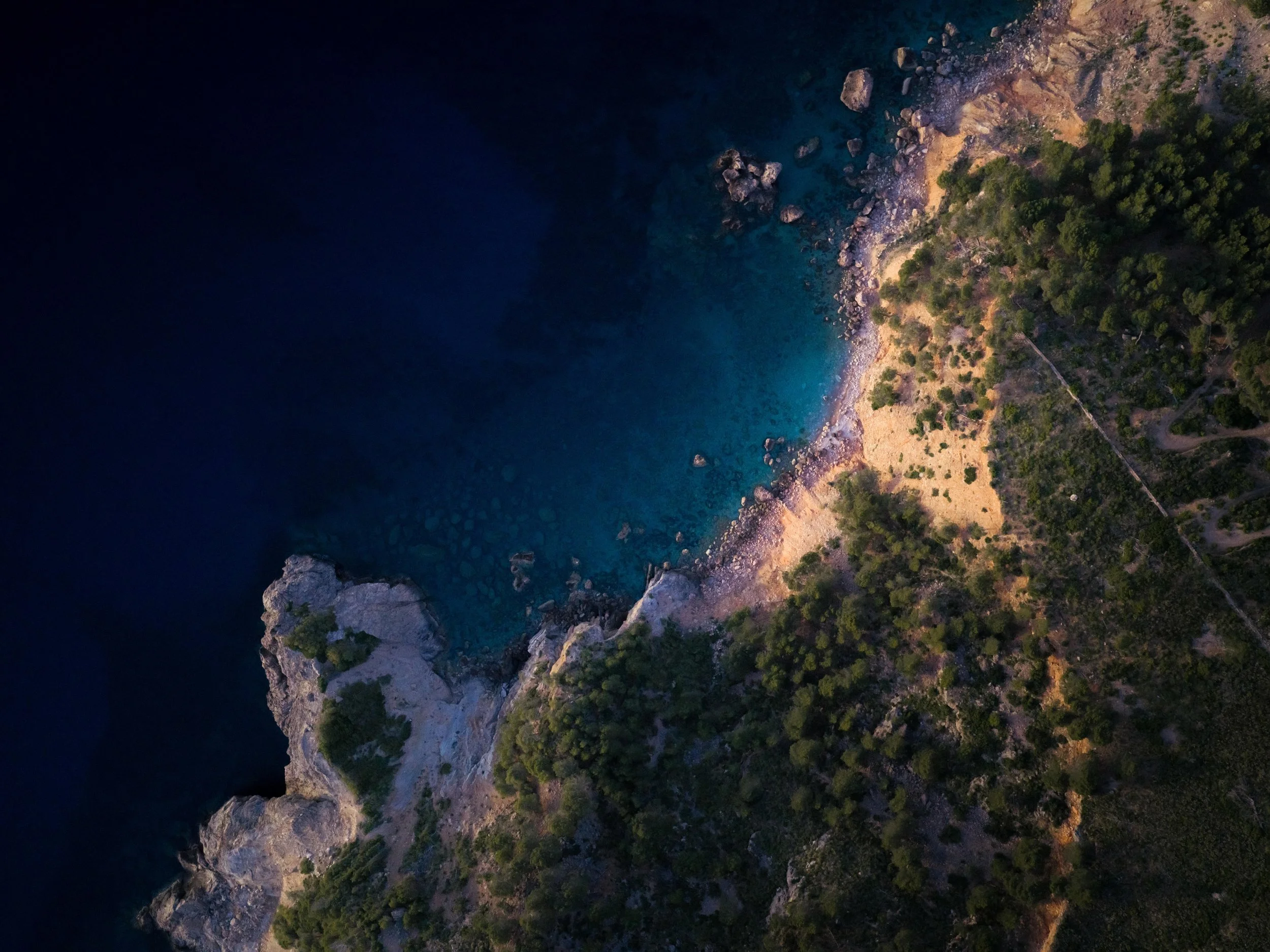 Aerial view of a rugged coastline with large rocks, sandy beaches, and green trees along the shore, taken at dusk or dawn.