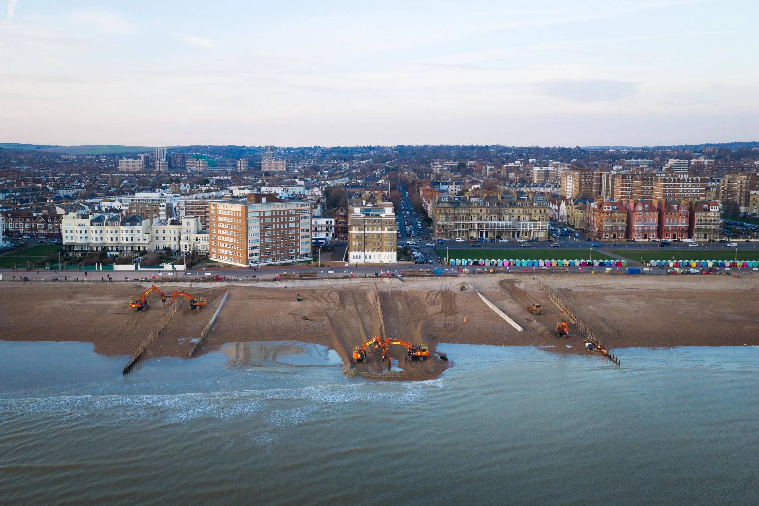 A cityscape view with tall buildings and a waterfront under a cloudy sky. In the foreground, construction equipment is working on a shoreline, with water and emerging land visible.