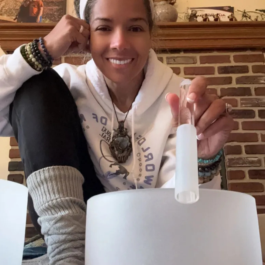 Woman smiling, wearing a hoodie, holding a plastic container with a white censor cap, sitting in a room with a brick fireplace and decorations on the mantel.