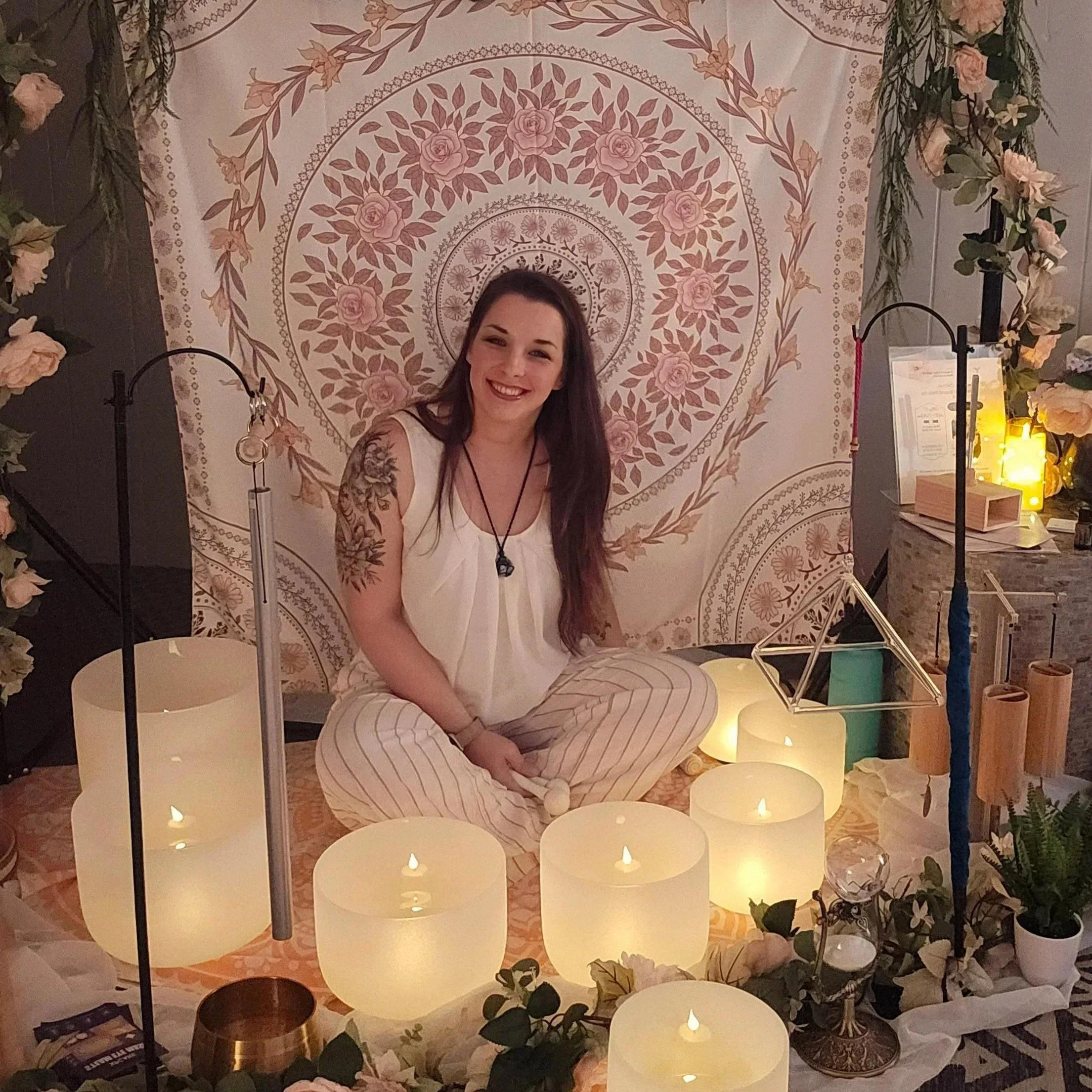 A woman with long dark hair and tattoos on her arm sitting cross-legged in front of a decorated backdrop with candles and flowers.