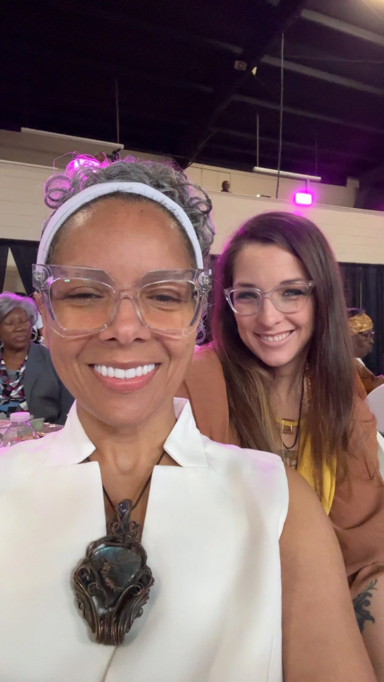 Two smiling women taking a selfie at an indoor event with other attendees in the background.