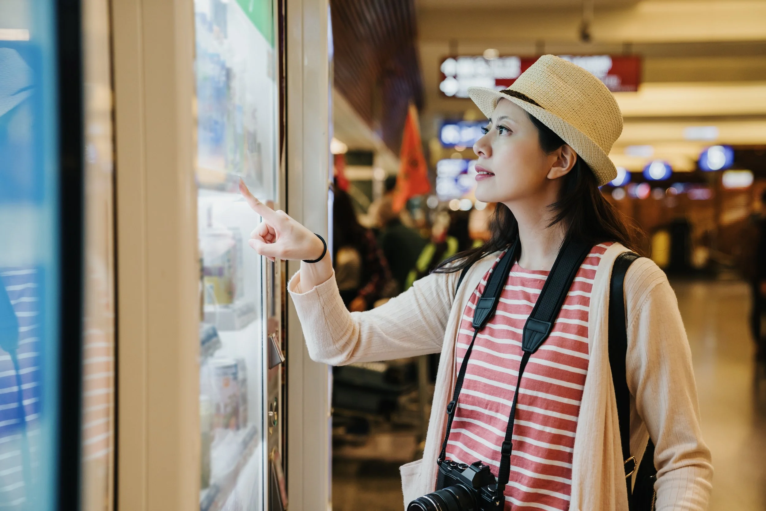 Young woman with a camera around her neck looking at vending machines in an airport or train station
