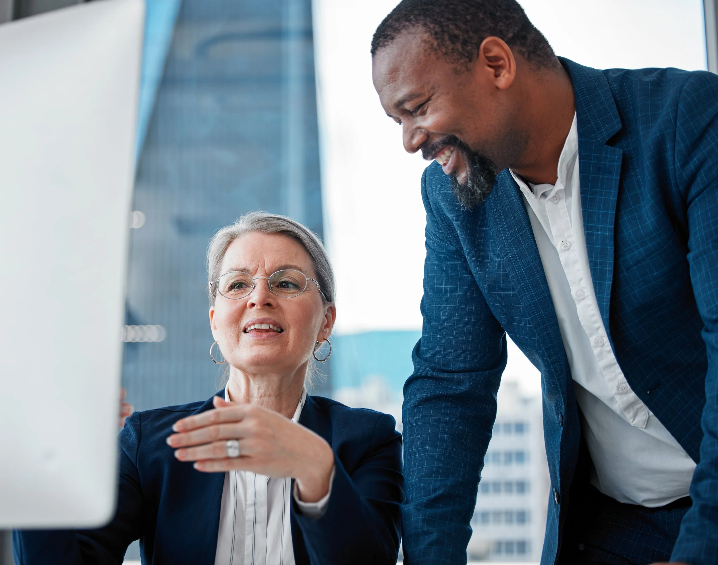 A woman with gray hair, glasses, and wearing a navy blazer and white shirt is seated, looking at her computer screen and speaking. A man with dark hair and beard, wearing a blue checked blazer and white shirt, is leaning over her shoulder, smiling and engaged in the conversation. They are in a modern office with large windows and city buildings in the background.