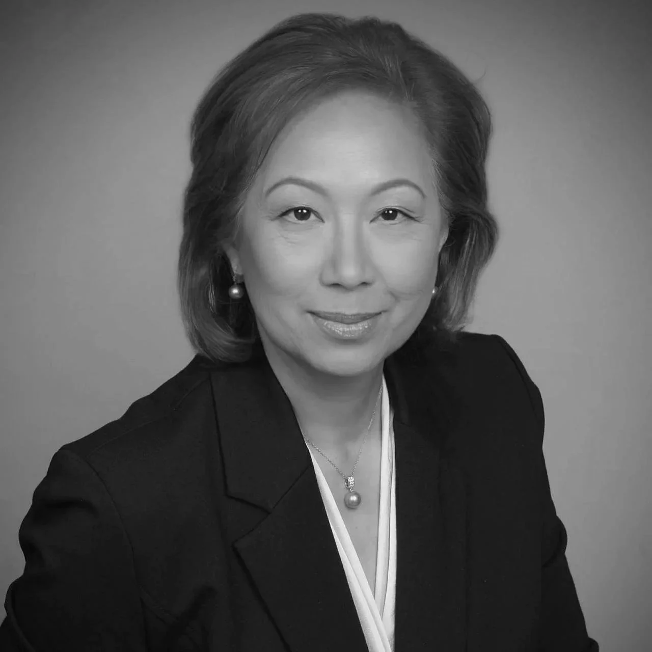 Black and white professional headshot of a woman with shoulder-length hair, wearing a dark blazer, pearl jewelry, and a confident expression.