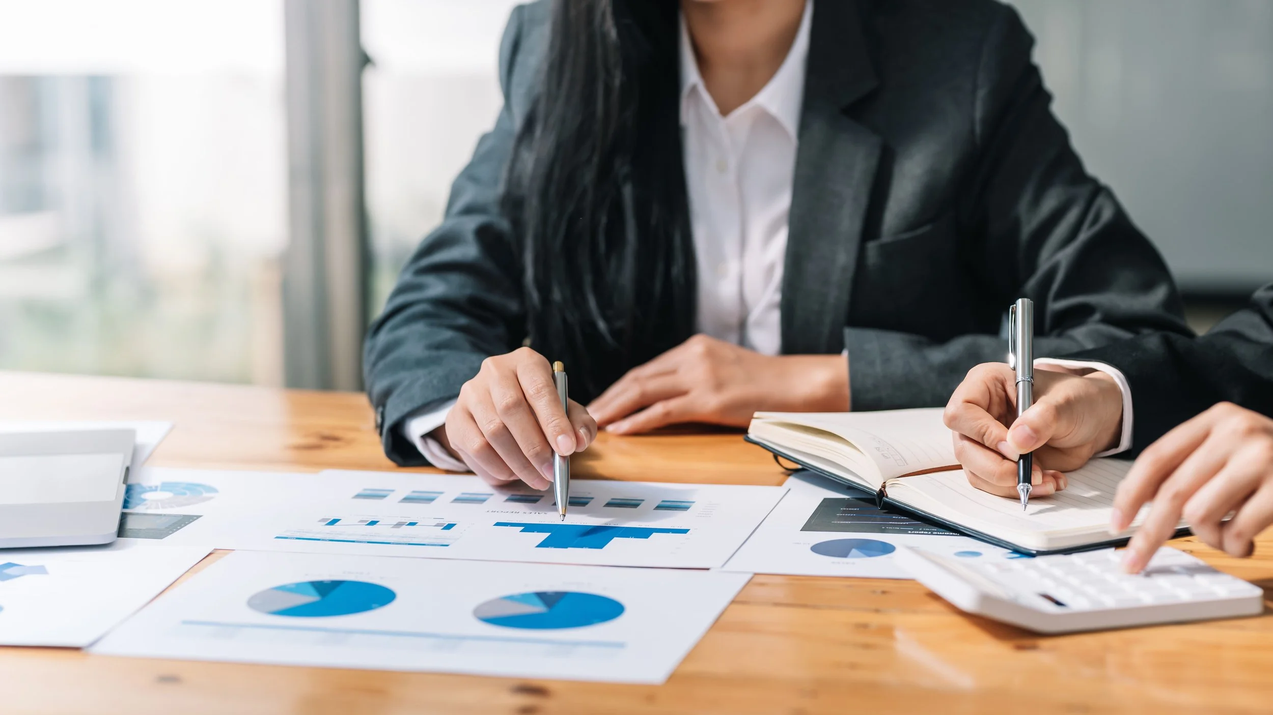 Businesspeople analyzing financial charts and documents on a wooden table, with notebooks and calculators.