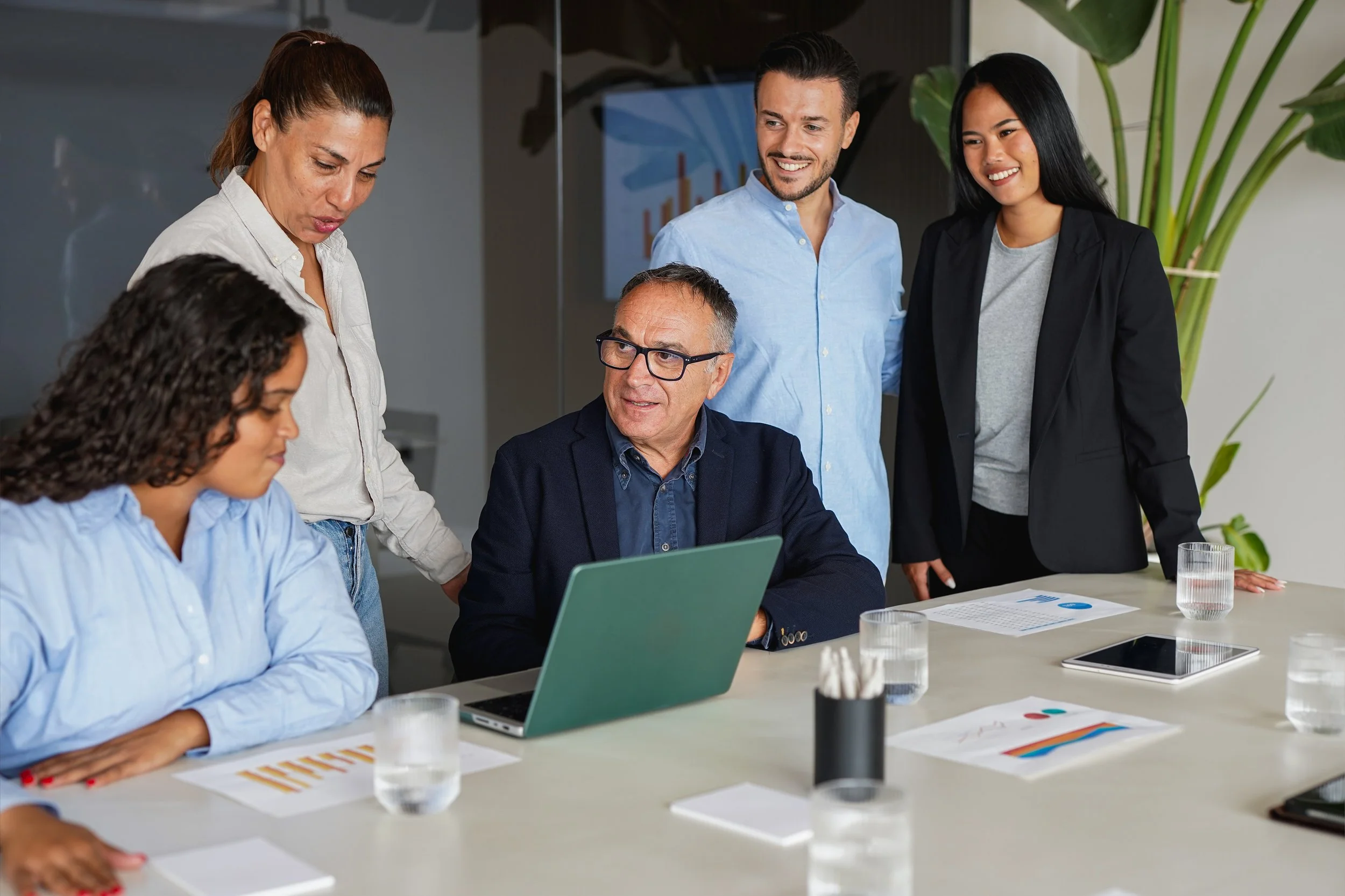 Five diverse professionals in a business meeting around a table, with documents, a laptop, and glasses of water, engaged in discussion in a modern conference room.