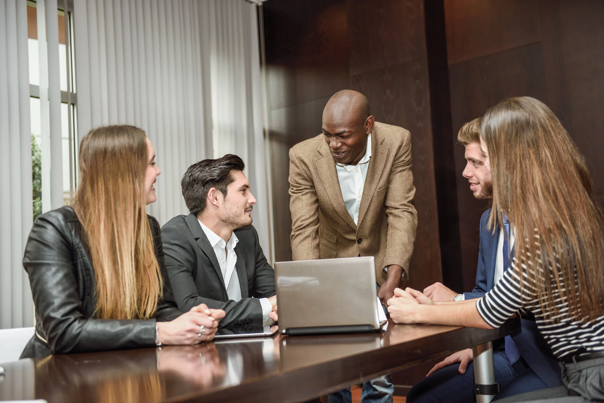 Five business professionals in a meeting room engaging in discussion around a table, with one standing and four seated, three men and two women, with a laptop open on the table.