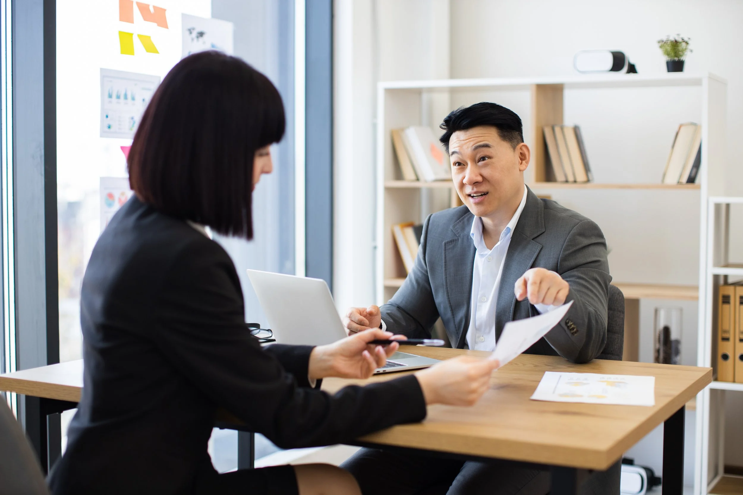 A man in a gray suit and a woman in a black blazer sitting at a table inside an office. They are engaged in a discussion, with the man smiling and gesturing towards the woman who is holding a paper and a pen. There is a laptop, some documents, a bookshelf, and some office decor in the background.