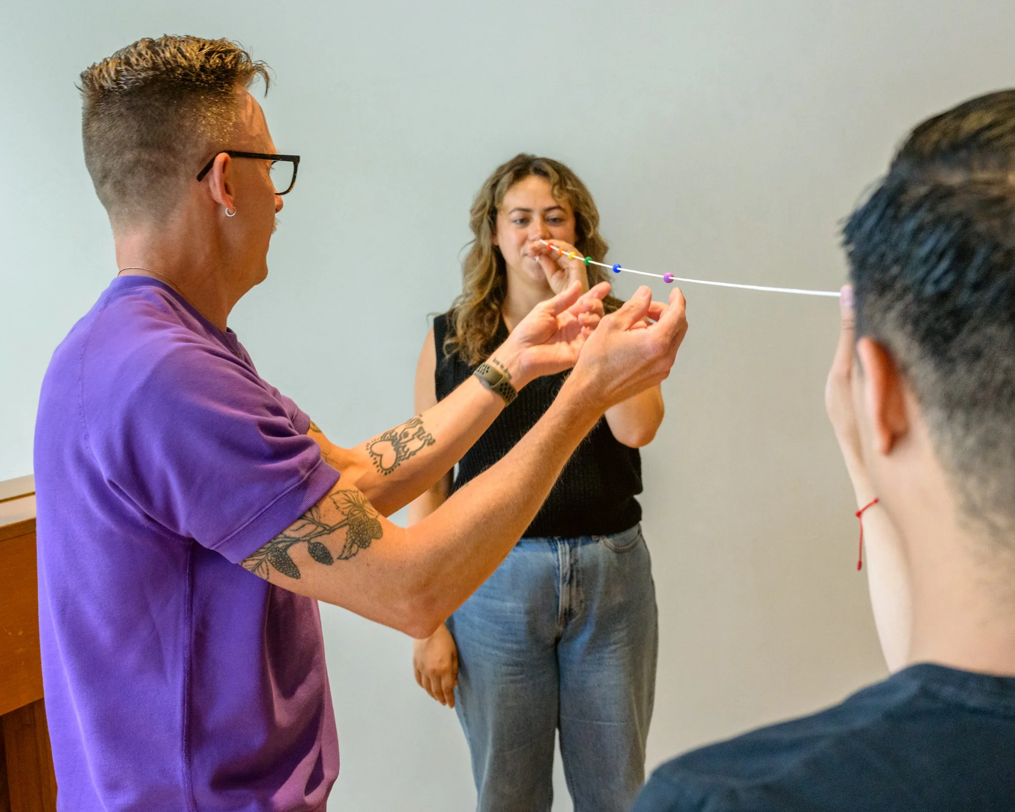 Tatiana and Jaryd each hold one end of a Brock String, a white string with colored beads on it. David stands in the middle, stabilizing the string.