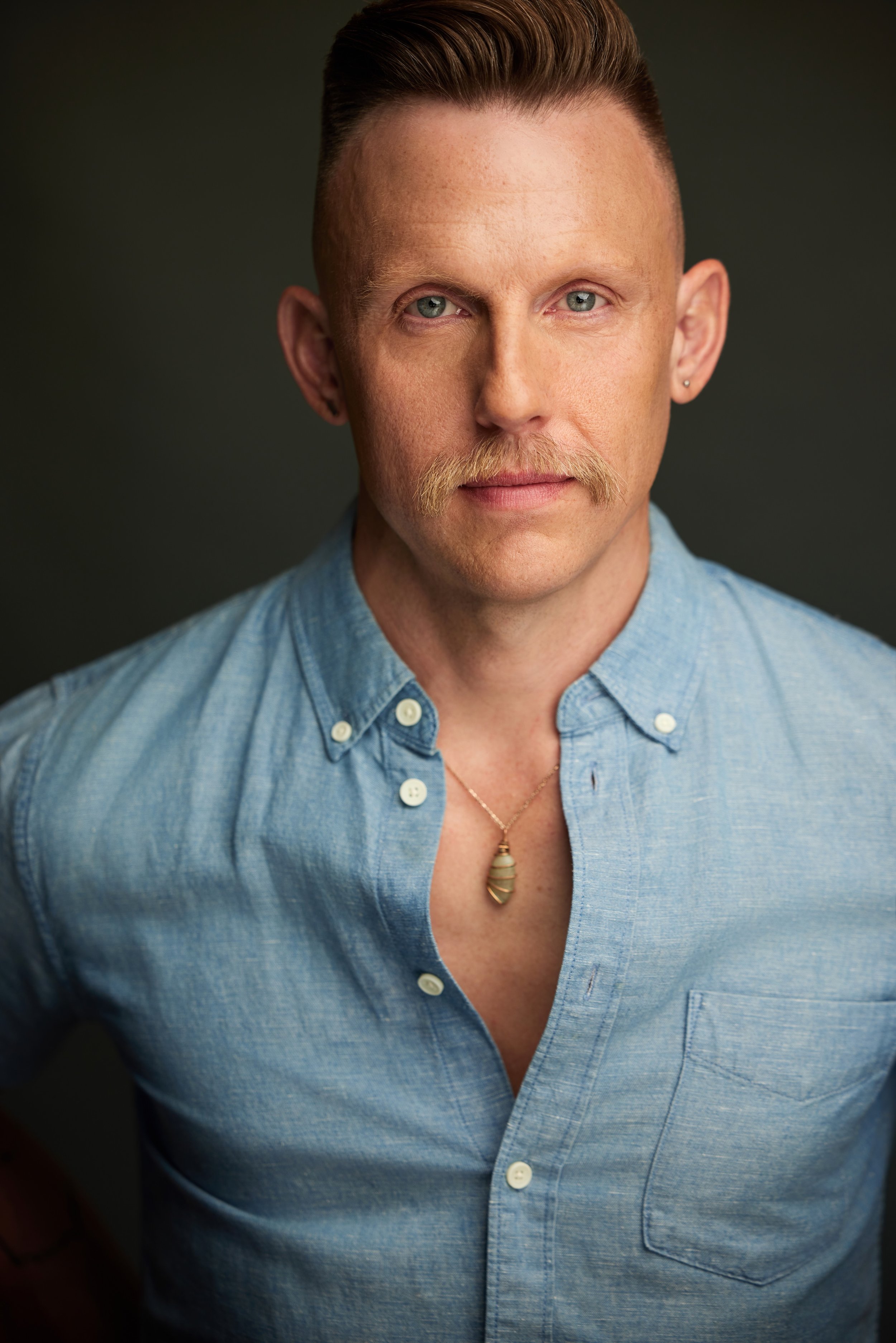 A man with light skin, short slicked-back hair, blue eyes, and a mustache, wearing a light blue denim shirt and a necklace with a pendant, against a dark background.
