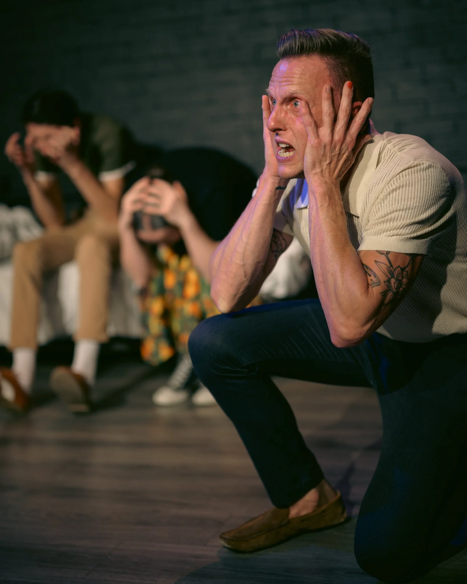 A man with a distressed expression, holding his head with both hands, sitting on the floor. In the background, another person with dark hair and glasses is also sitting, leaning forward with hands covering their face.