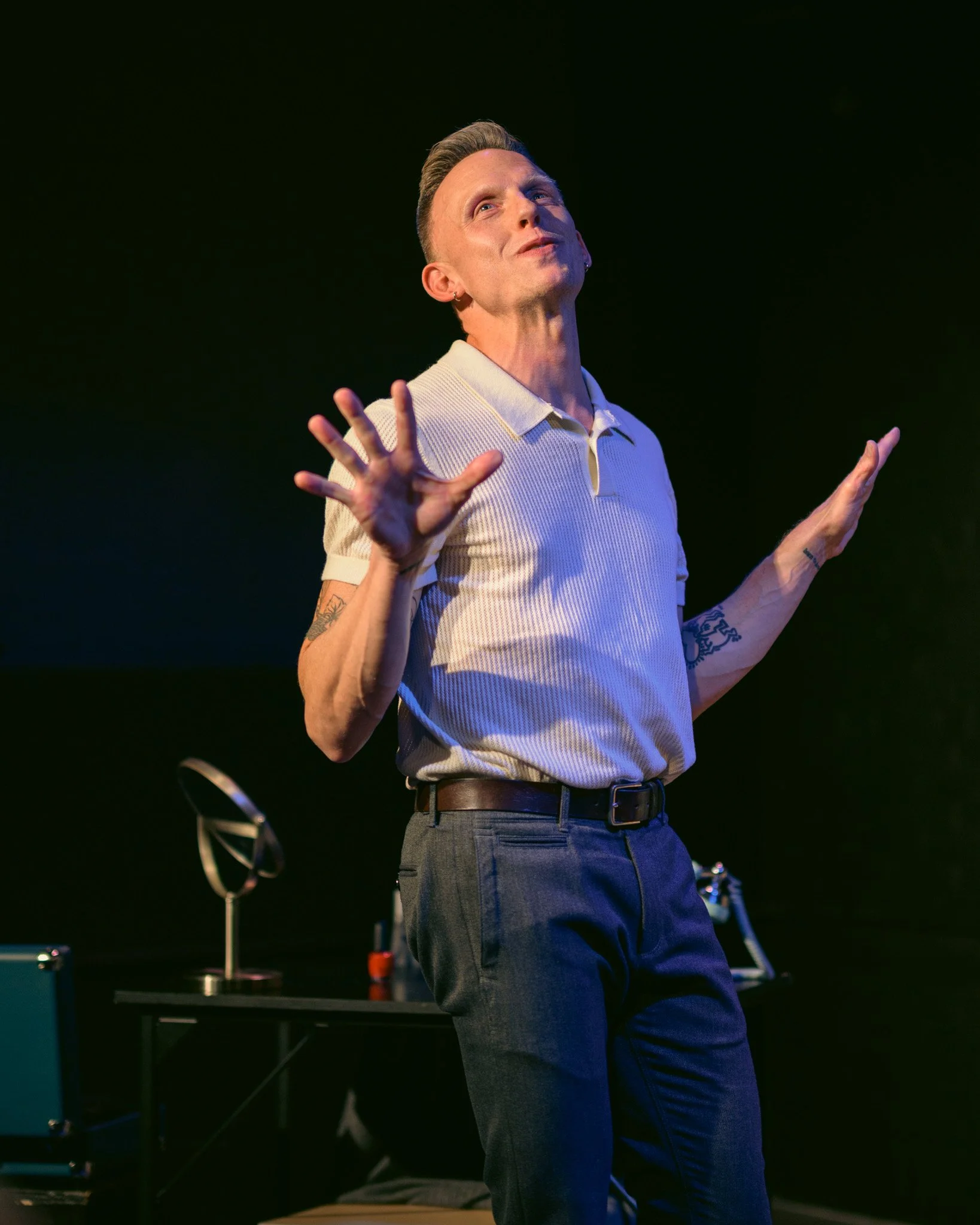 A man with short, styled hair and earrings, wearing a light-colored polo shirt and gray pants, standing on a stage with a dark background. He has tattoos on his arms and is gesturing with his hands while speaking, with a confident and expressive faci