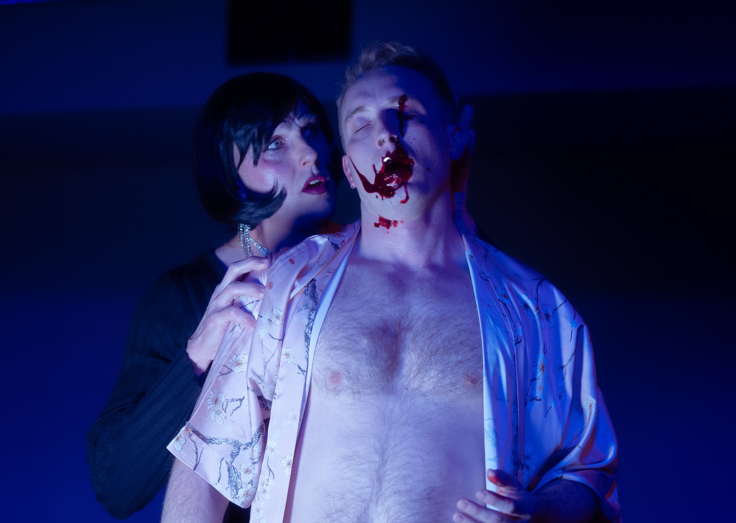 A man with blood on his face and chest, kneeling with his eyes closed, wearing a silk robe, while a woman with dark hair and earrings is behind him, touching his neck in a dark blue-lit scene.