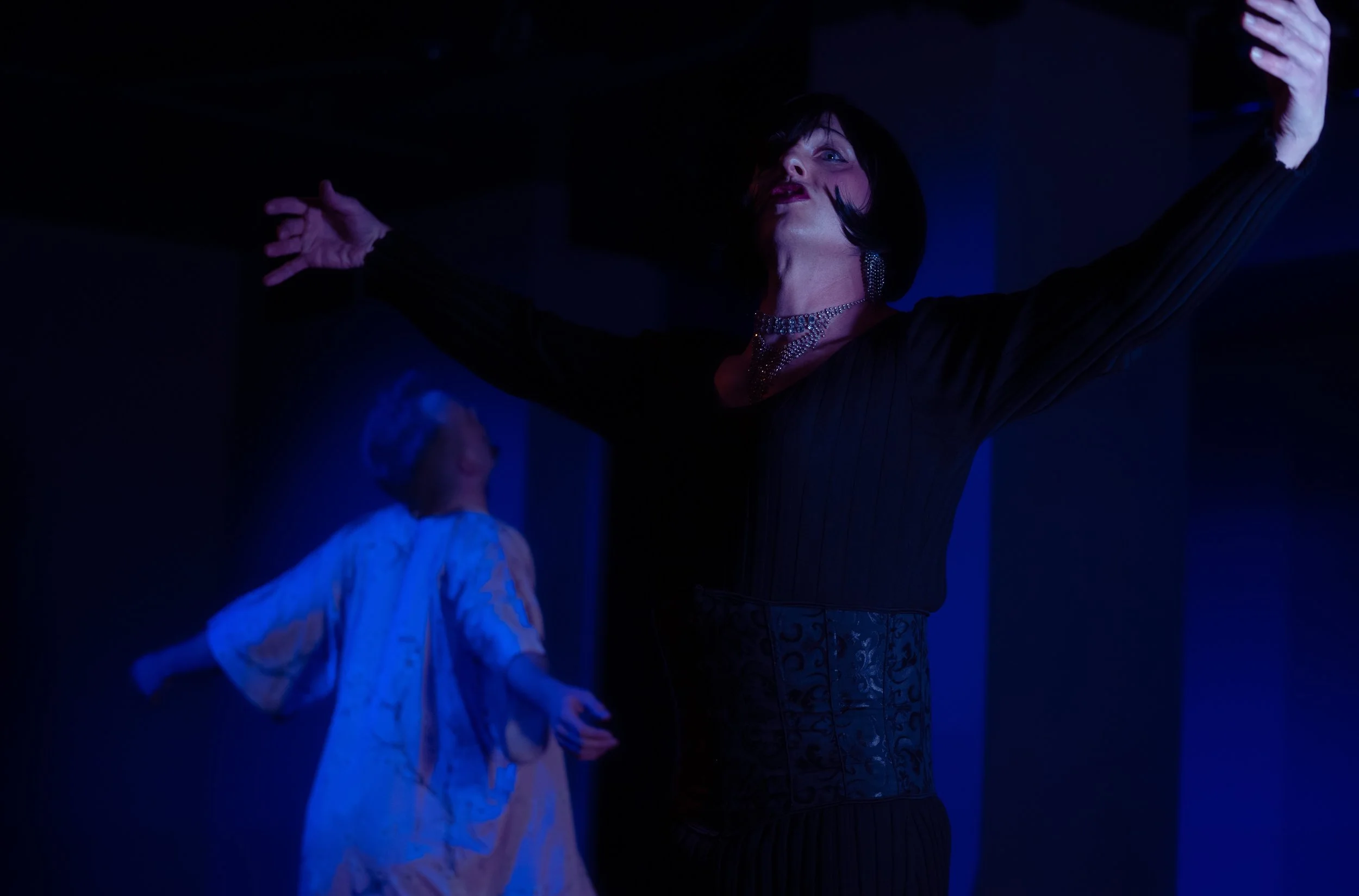 A woman with short dark hair wearing a black dress and rhinestone necklace dancing with her arms raised in a dark, blue-lit nightclub. A person in a white lace top is visible in the background.