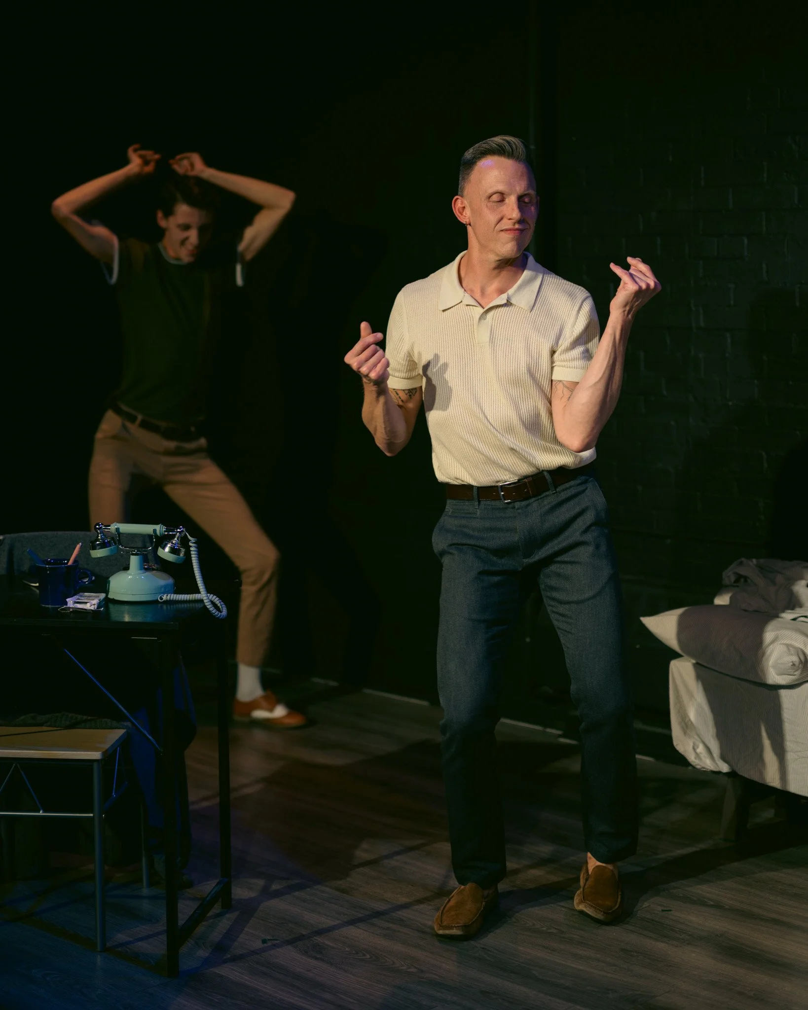 A man wearing a cream polo shirt and gray pants dancing with eyes closed, gesturing with his right hand, in a dimly lit room with a woman in the background in black attire, tying her hair back.