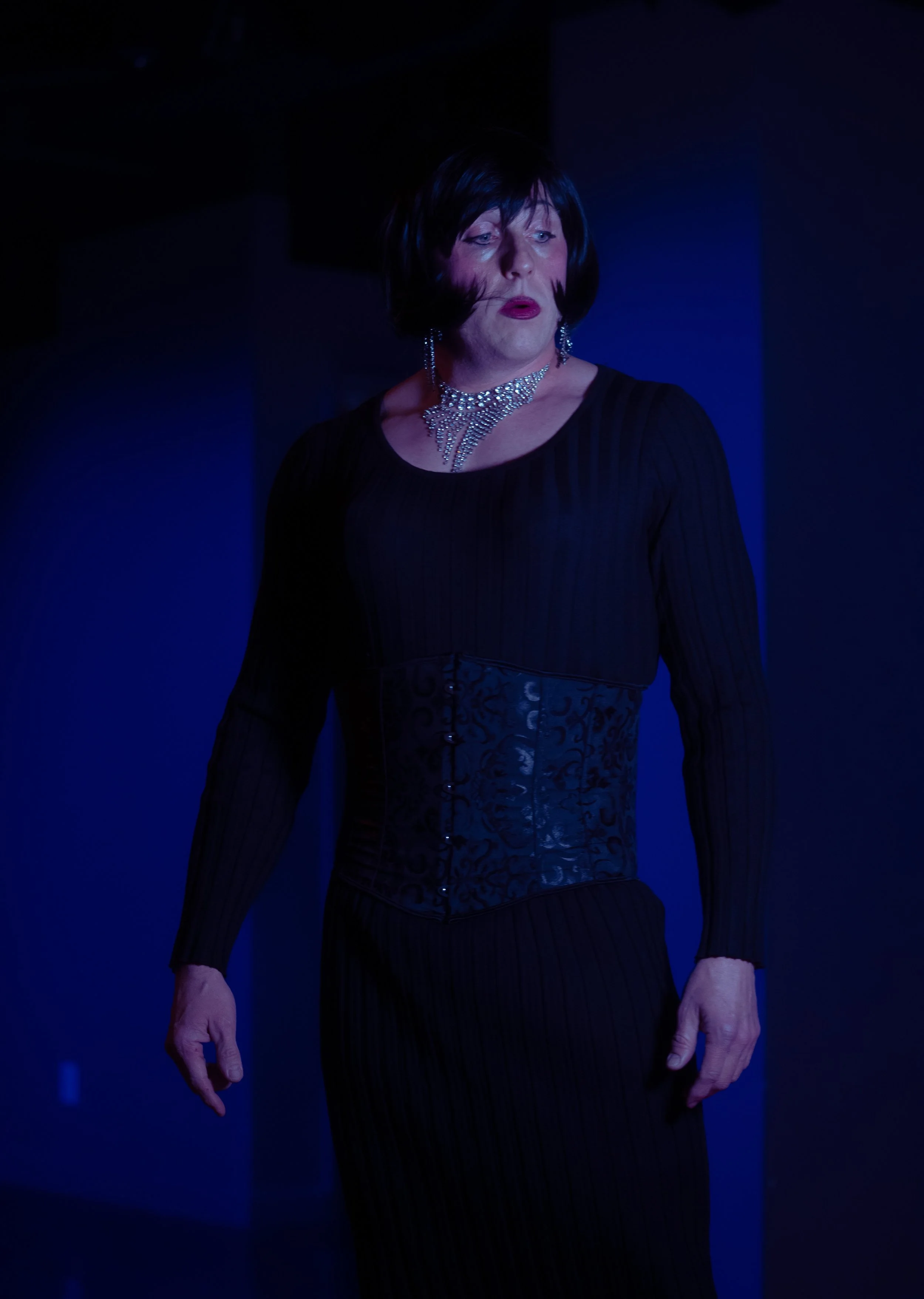 A person dressed in dark, elegant clothing with jewelry including a necklace and earrings, standing in a dimly lit environment with blue lighting.