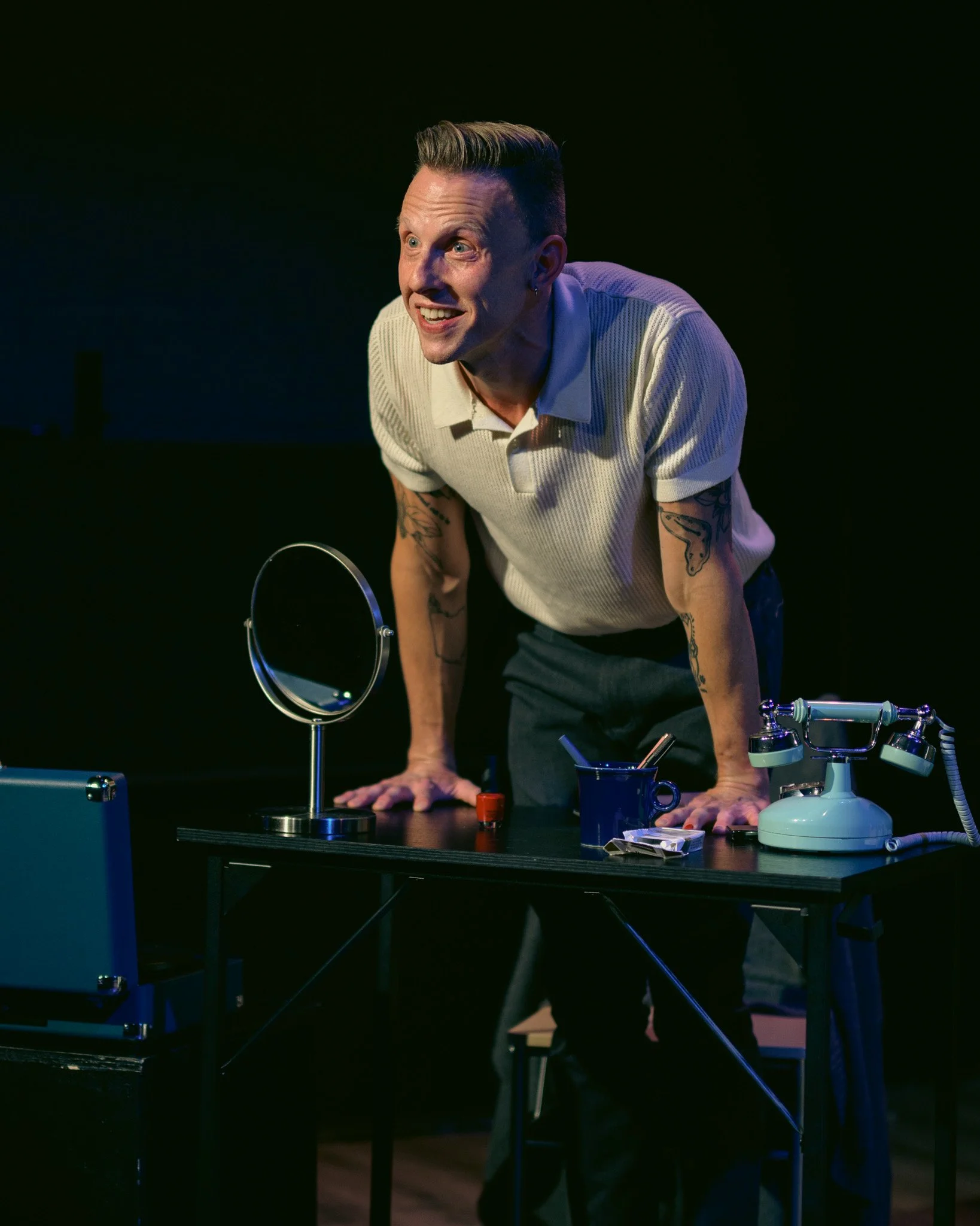 A man with short hair and tattoos on his arms leaning forward on a black table with various objects, including a mirror, a mug, and a vintage telephone, in a dark room with a black background.