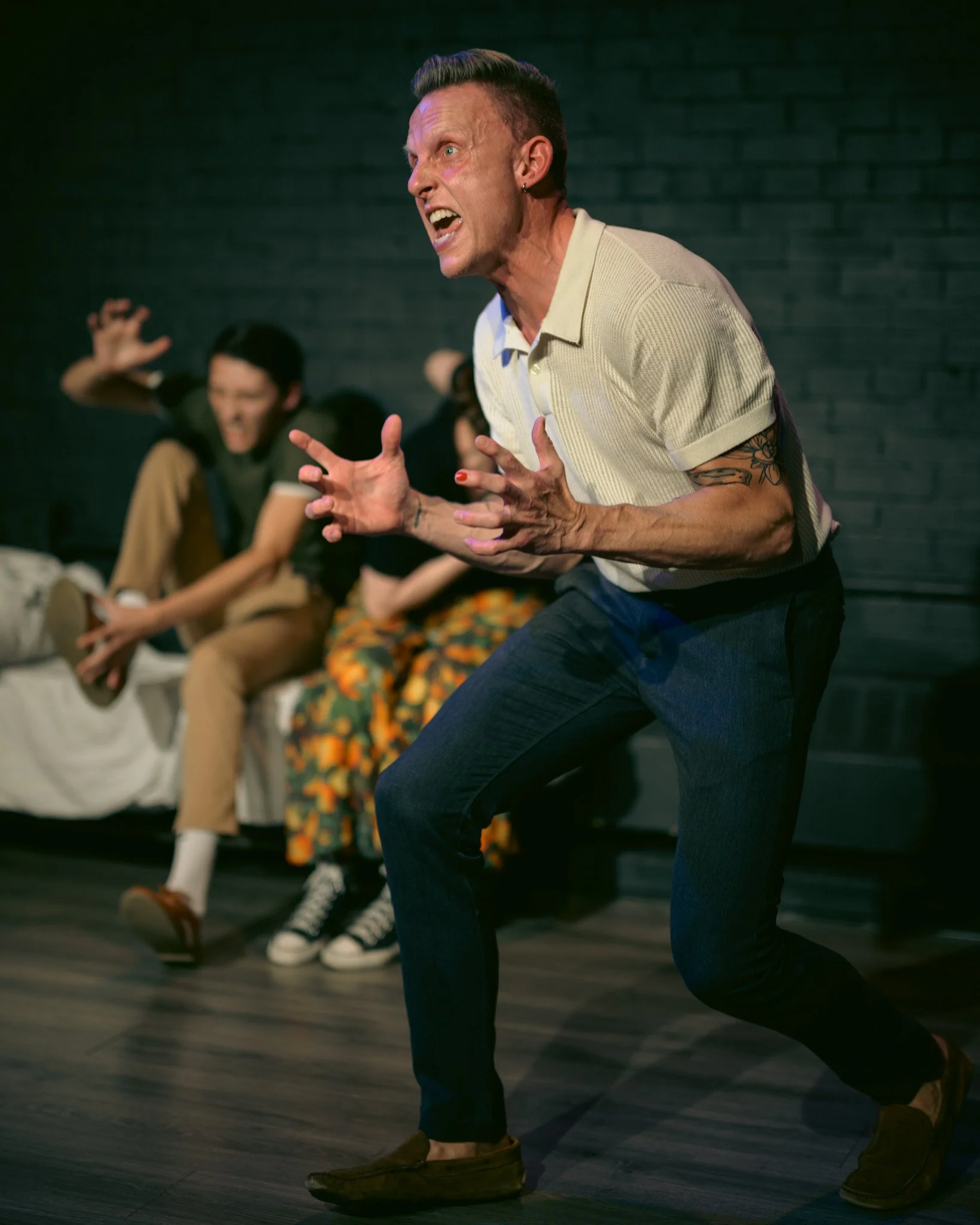A person passionately acting or shouting during a theatrical performance, with other seated individuals in the background on a stage with a dark brick wall.