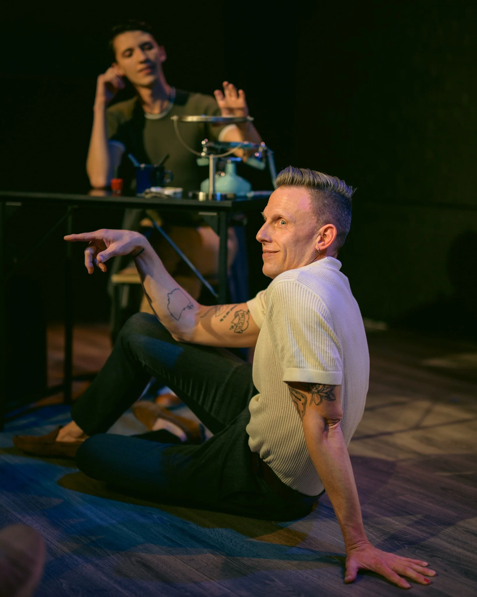 Two performers engaging in a theatrical or dance performance on stage with dark background. The person in the foreground is sitting on the floor, supporting themselves with one arm, has short hair with a fade haircut, tattoos on their arm, and is gaz