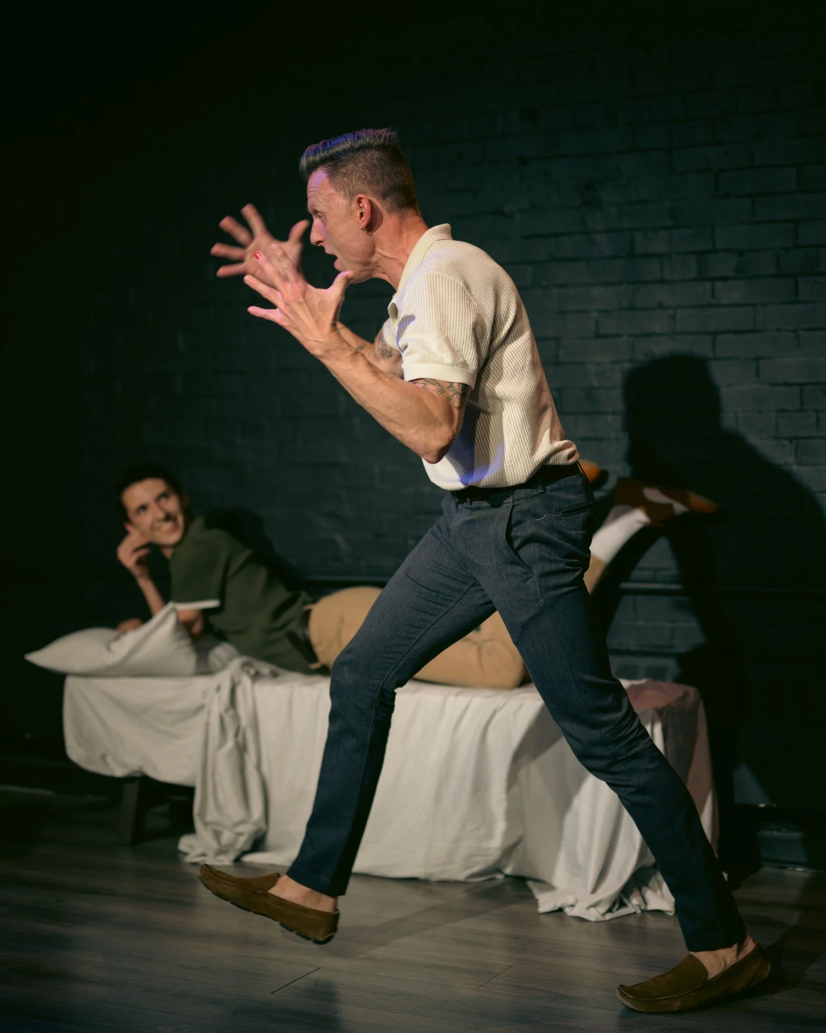 Man dressed in retro style with a pompadour hairstyle, wearing a beige polo shirt, navy blue pants, and brown shoes, performing on stage with a black brick wall background. A woman on a bed in the background looks amused.