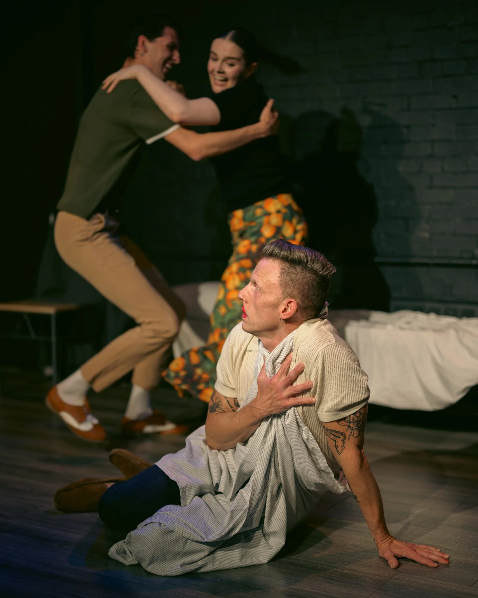 Group of four people engaging in dramatic play, with two standing and two sitting on the floor, two of them smiling, one with hand on chest, and the others gesturing with expressions of surprise or excitement, set against a dark brick wall background