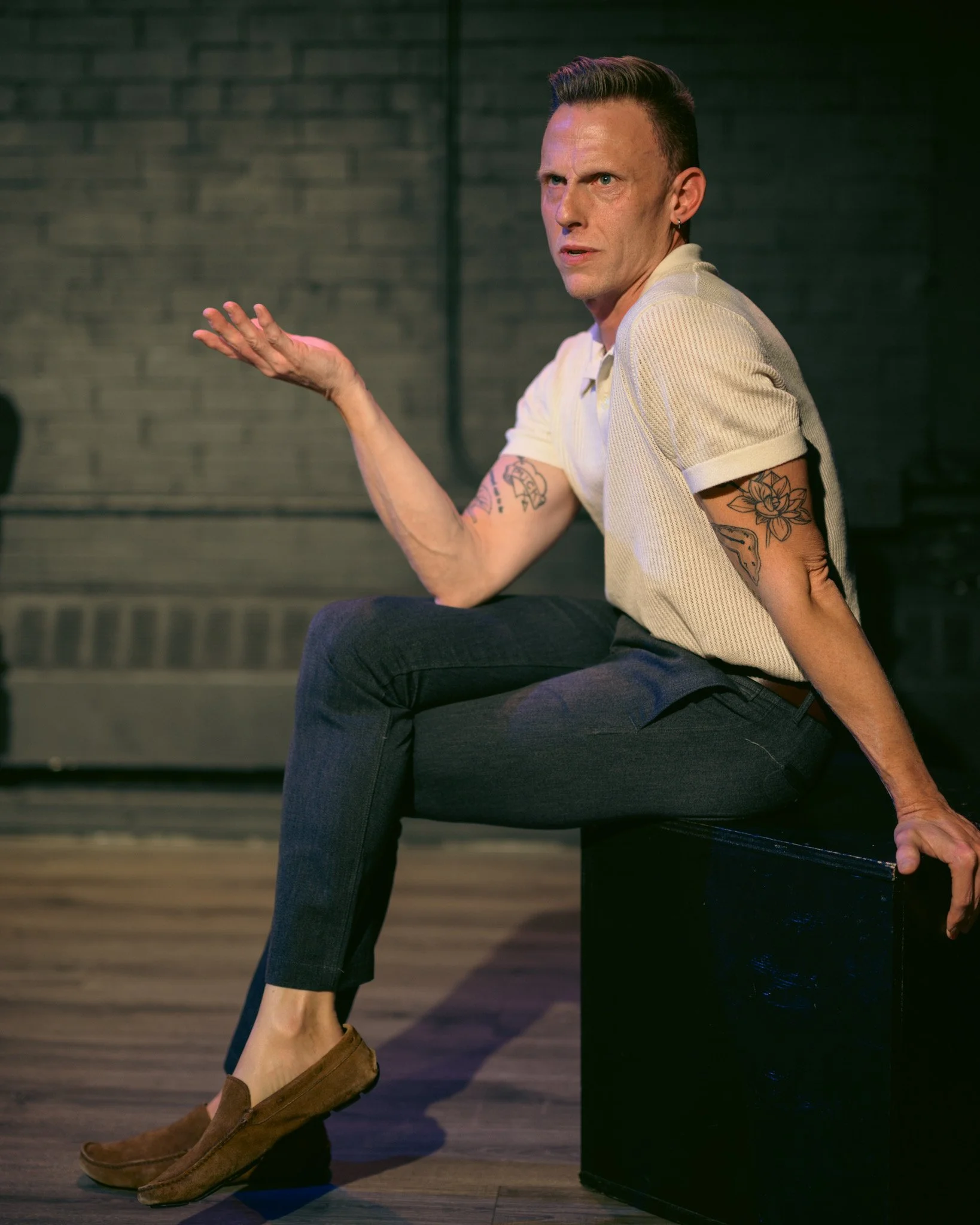 A man with tattoos wearing a yellow shirt and dark pants, sitting on a black box, gesturing with his right hand, in a dimly lit room with a brick wall background.