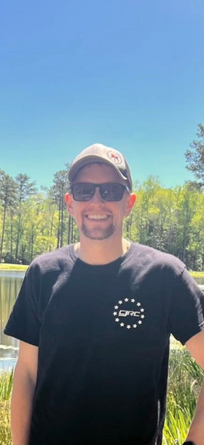 Smiling man wearing sunglasses and a baseball cap outdoors by a lake with trees and a clear blue sky