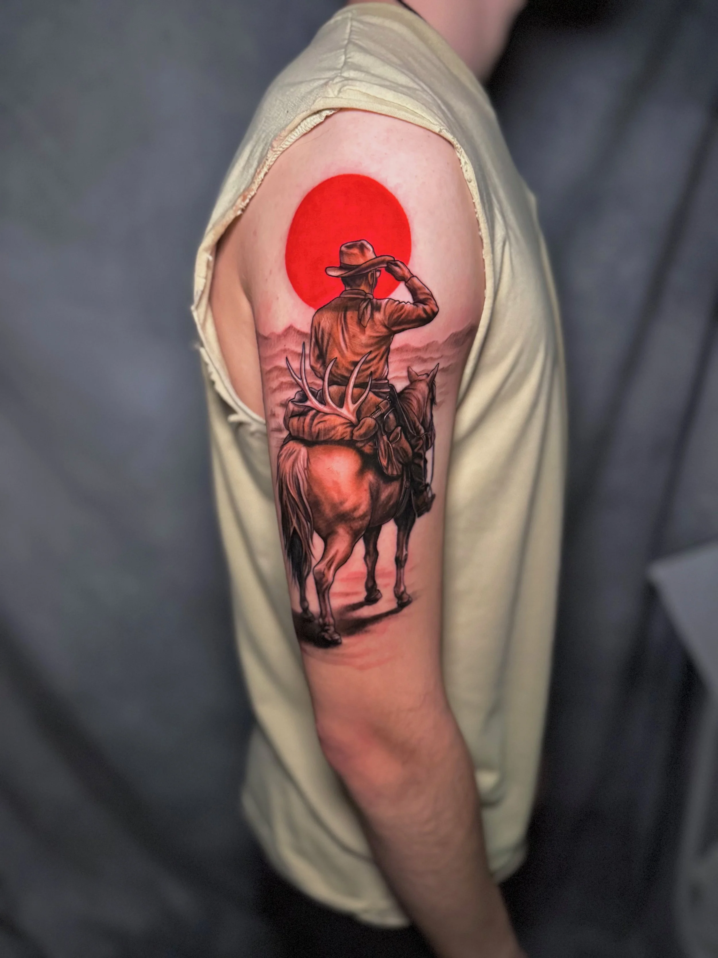 Tattoo of a cowboy riding a horse, with a large red sun behind him and mountains in the background.