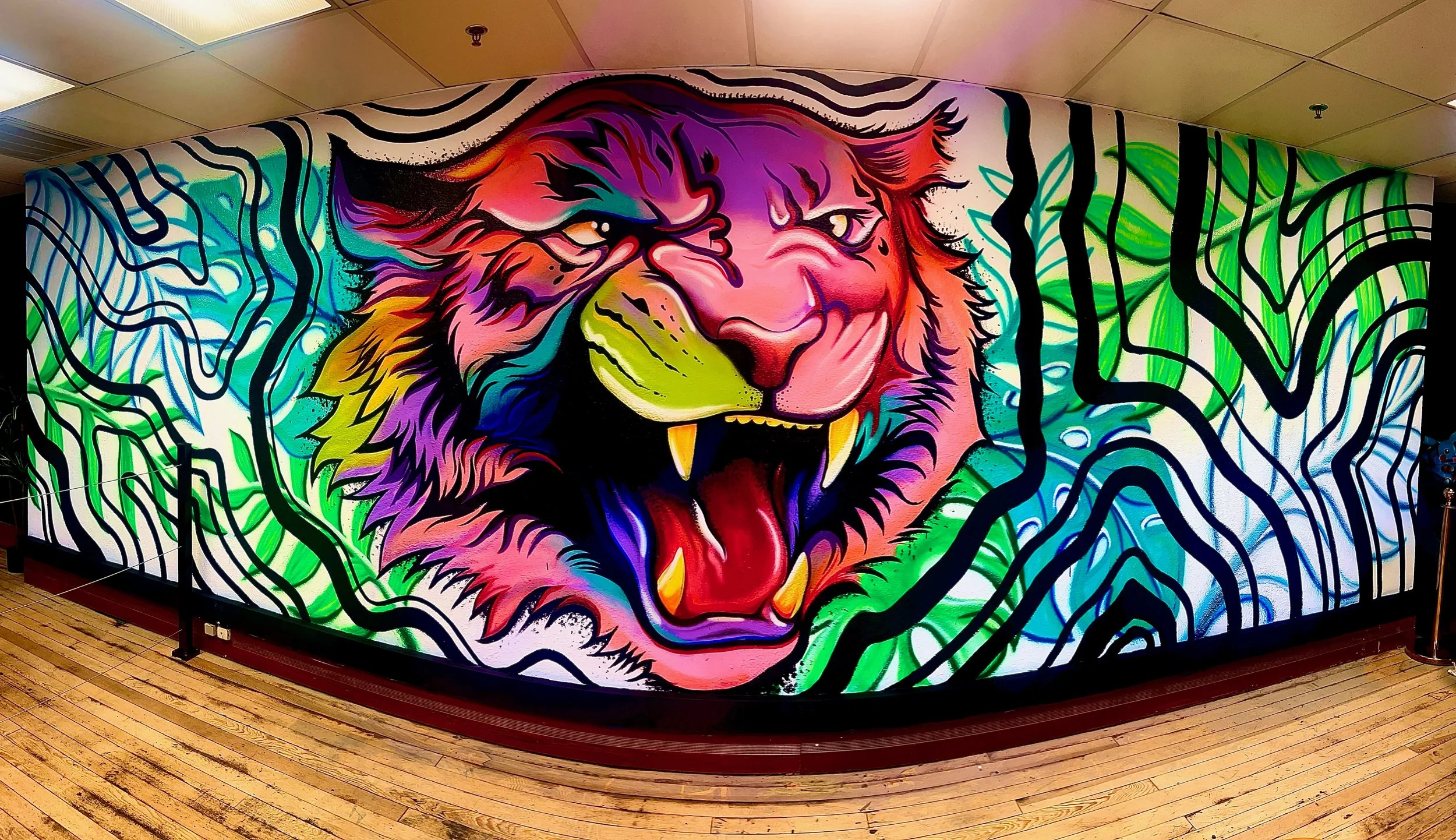 Colorful mural of a roaring tiger with an open mouth, yellow teeth, pink tongue, surrounded by abstract green, blue, black, and purple lines on a wall in an indoor space with wooden flooring.