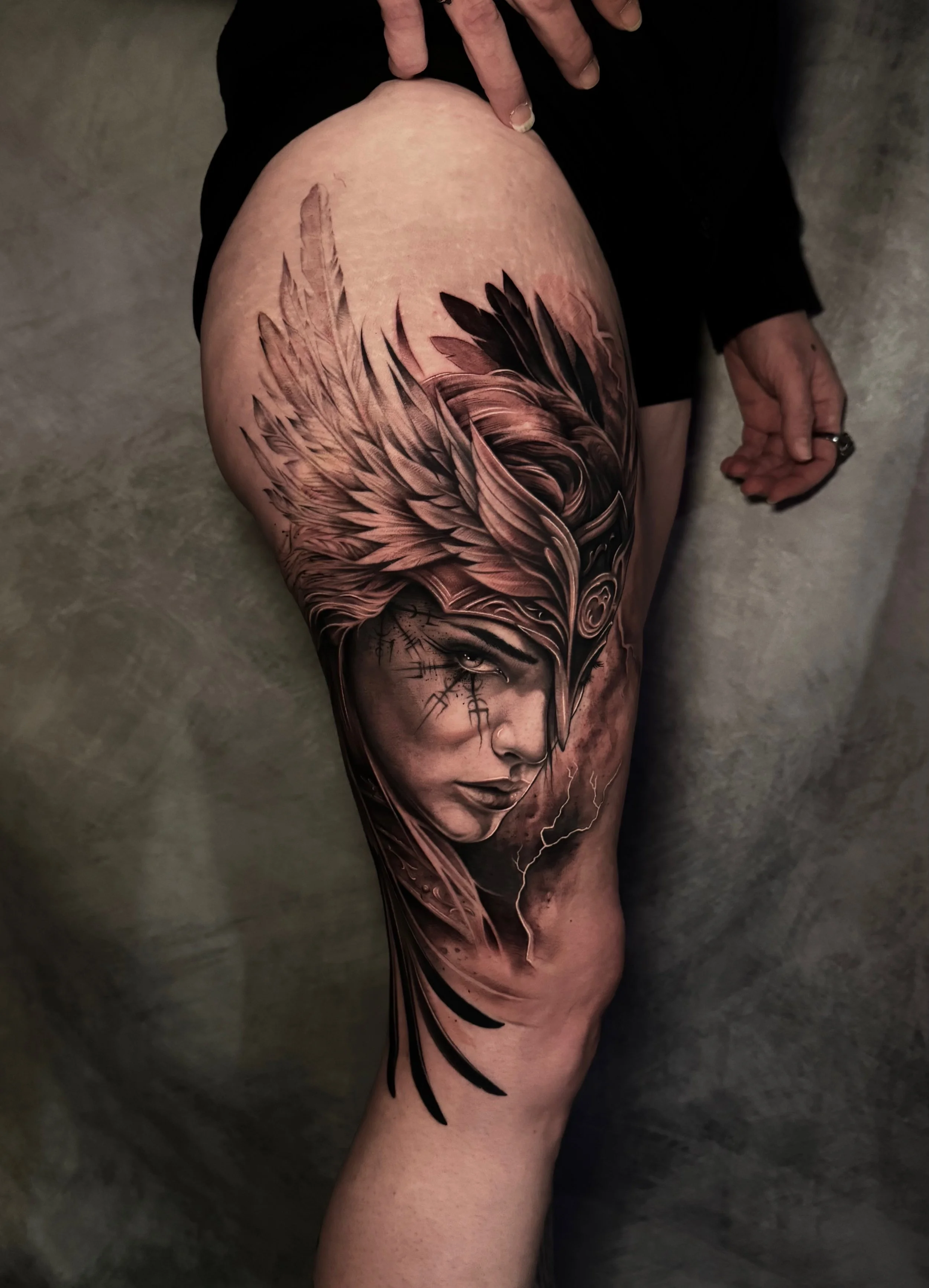 A detailed black and gray tattoo of a woman's face with a headdress made of feathers adorns a person's thigh.