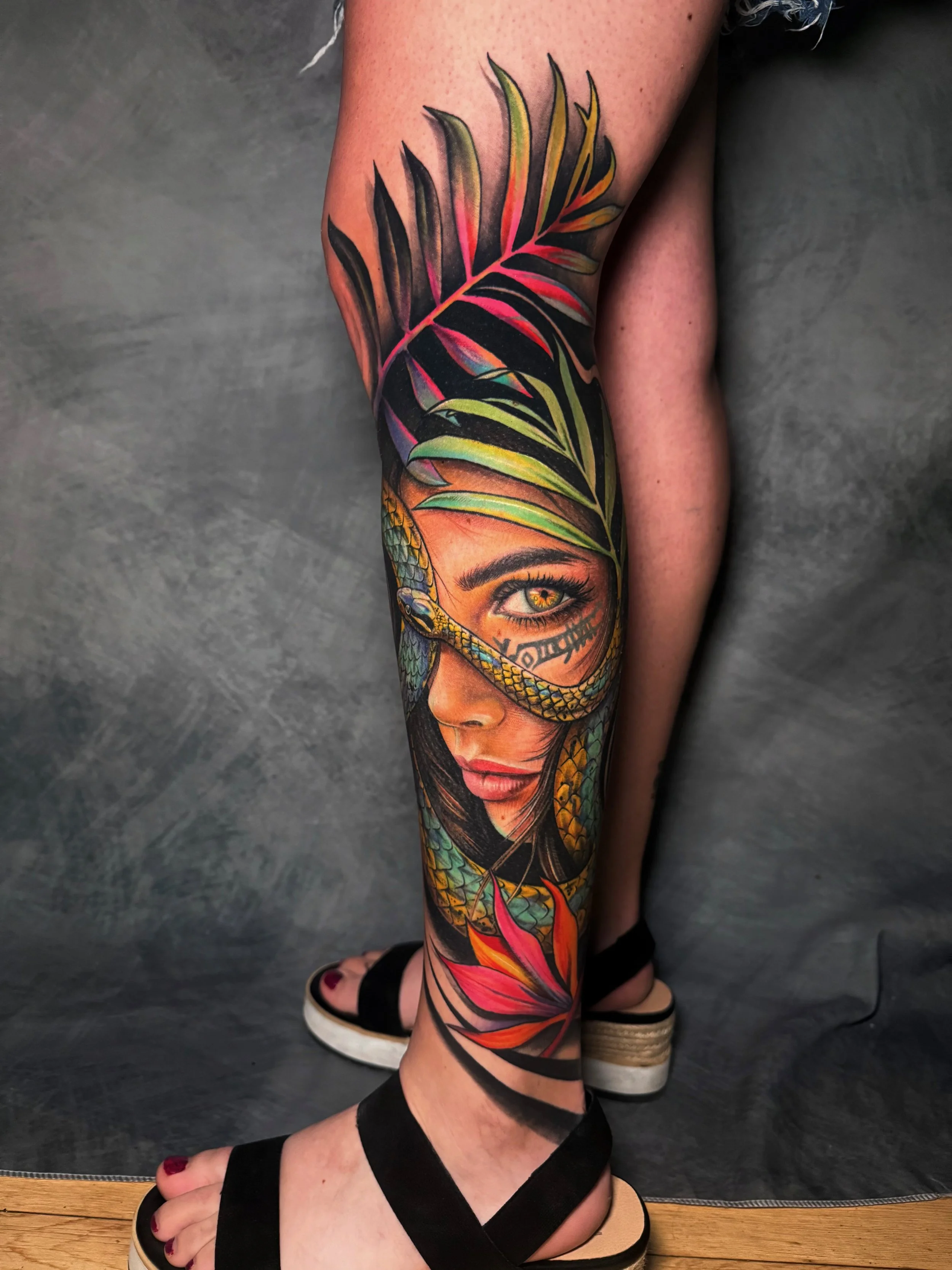 Colorful tattoo of a woman's face with a snake around her eye, surrounded by tropical leaves and flowers on her leg.