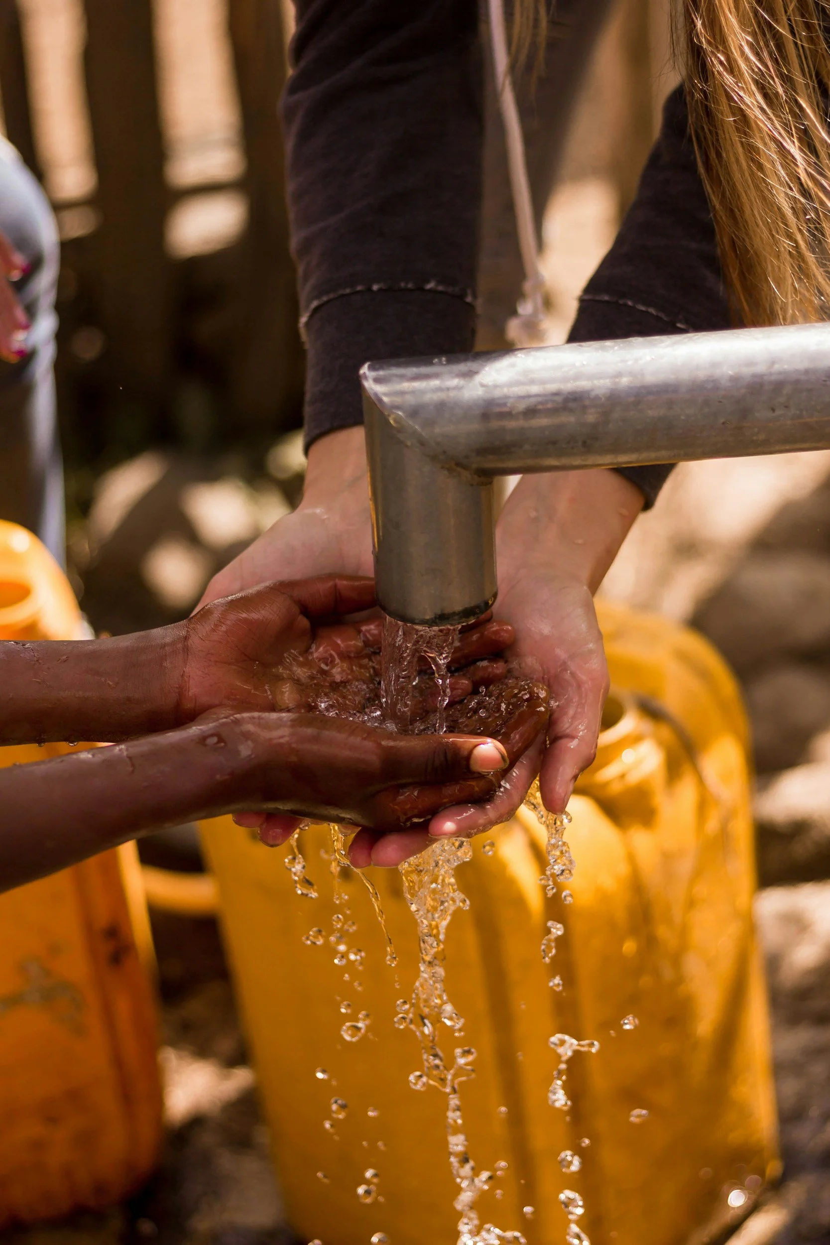 Two people washing hands under a running faucet outdoors, with yellow containers in the background.