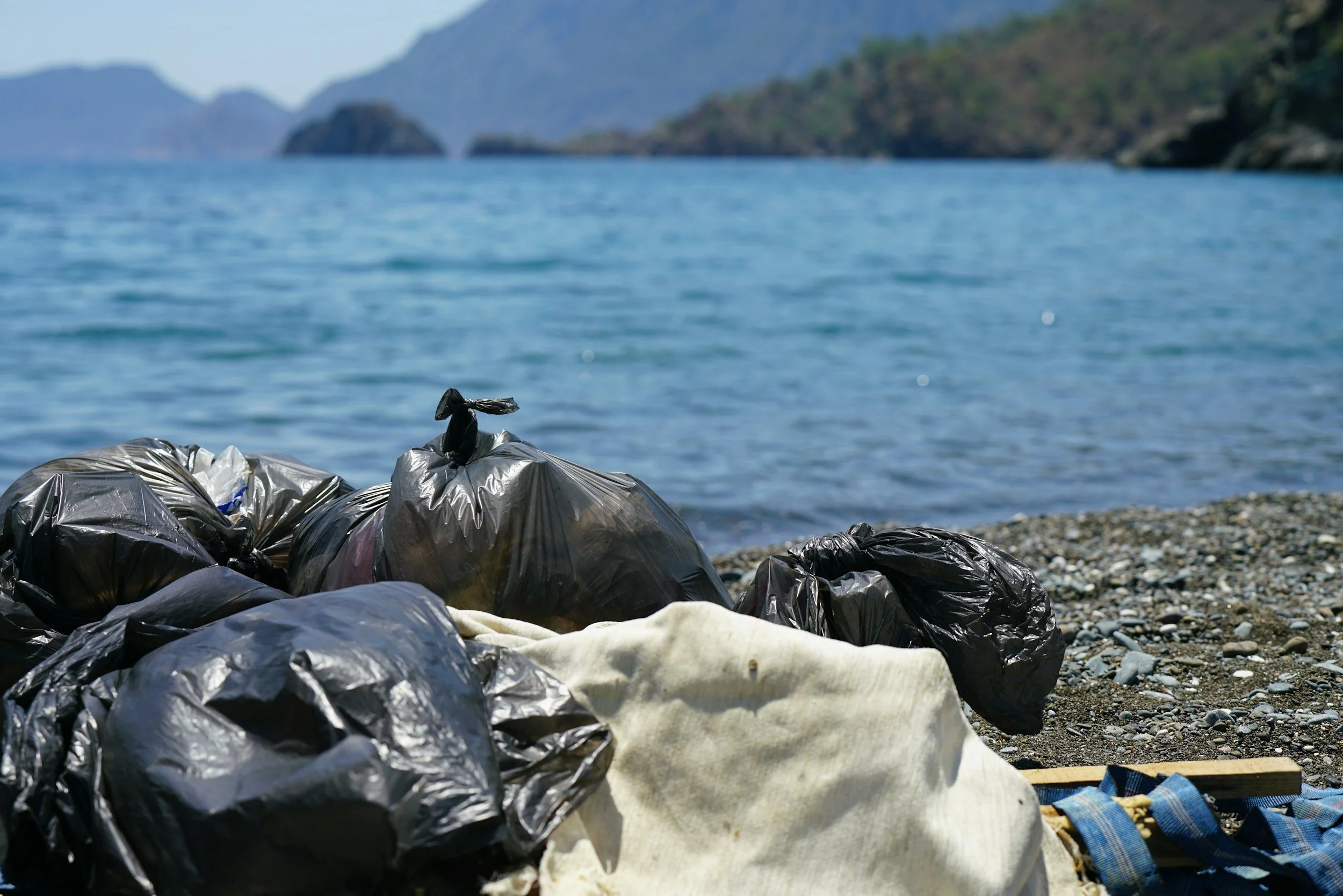 Bags of trash collected along a rocky shoreline with a blue lake and hills in the background.