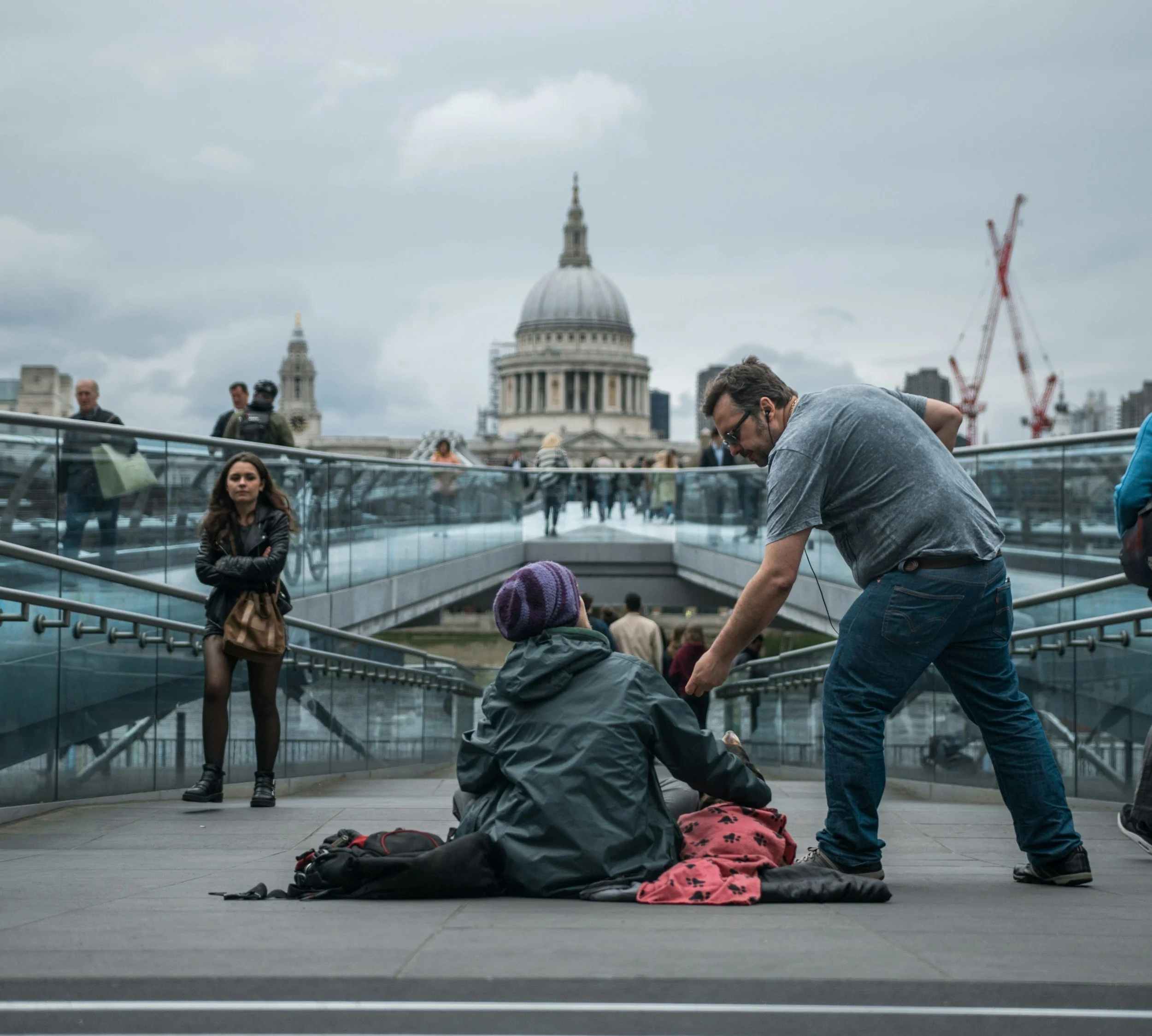 Two people sitting on the ground talking, one man standing talking to them, on a bridge with St. Paul’s Cathedral in London in the background, with other pedestrians walking by and construction cranes in the distance.