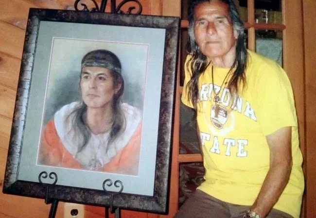 A man with long dark hair wearing a yellow Georgia State T-shirt sitting next to a framed portrait of a woman with long dark hair and a headband.