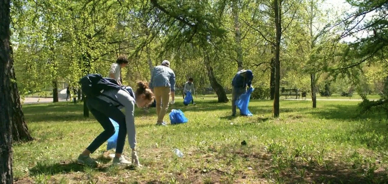 Group of people picking up trash in a park with trees and green grass.