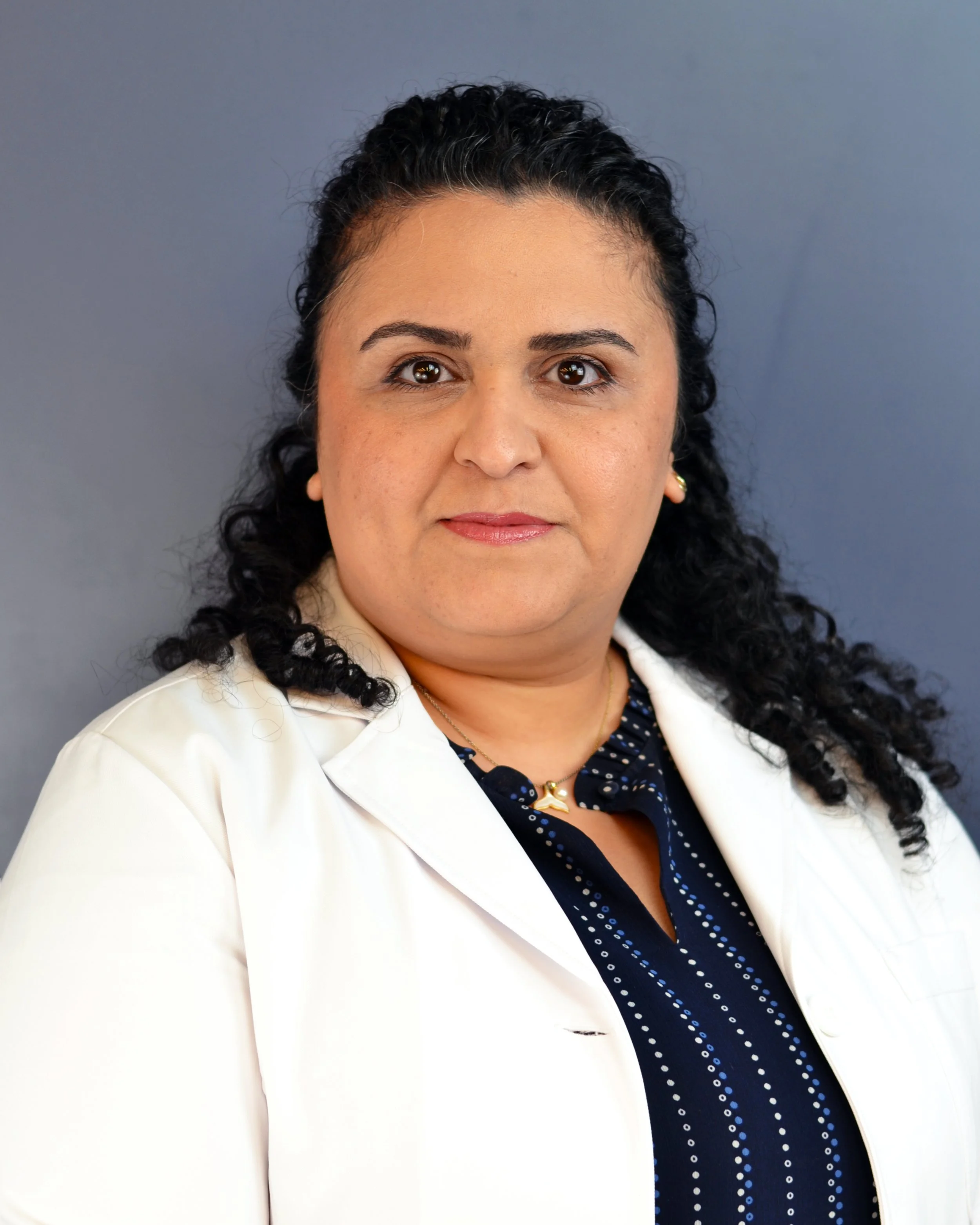 Professional headshot of a woman with dark curly hair, wearing a white blazer over a navy blue polka dot blouse, against a plain background.