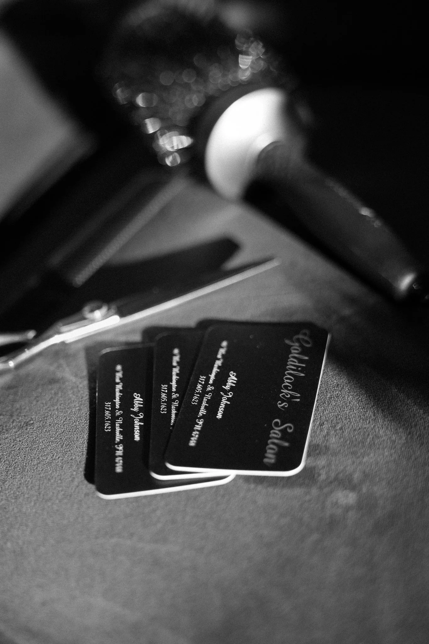 Black business cards with white cursive text reading "Hadioli's Salon" and contact information, placed on a textured surface. Part of a salon tool and a spray bottle are visible in the blurred background.