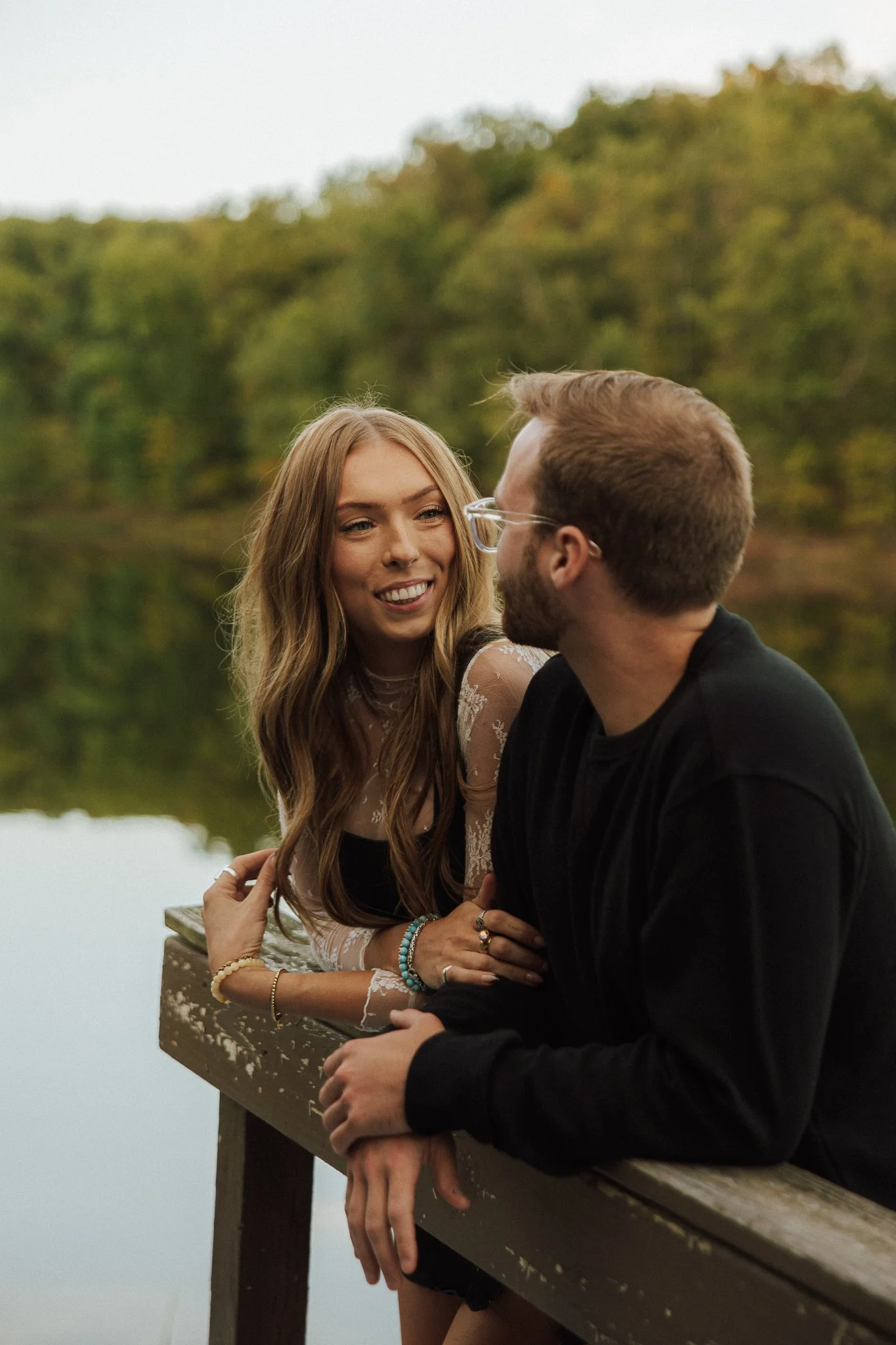A young woman and a young man are leaning on a wooden railing by a lake with trees in the background during daytime. They are smiling and looking at each other.'}