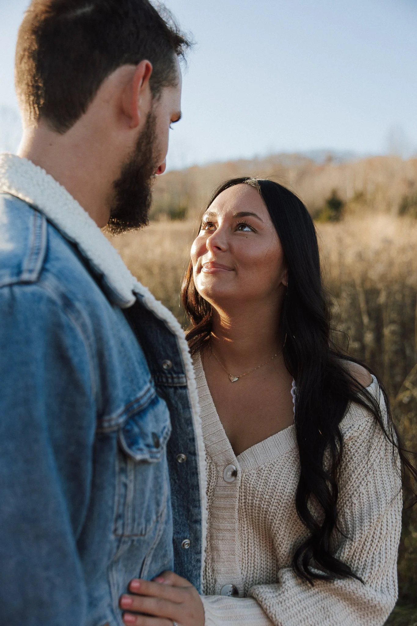 A man and woman standing outdoors, facing each other and looking into each other's eyes, with a blurred natural landscape in the background.