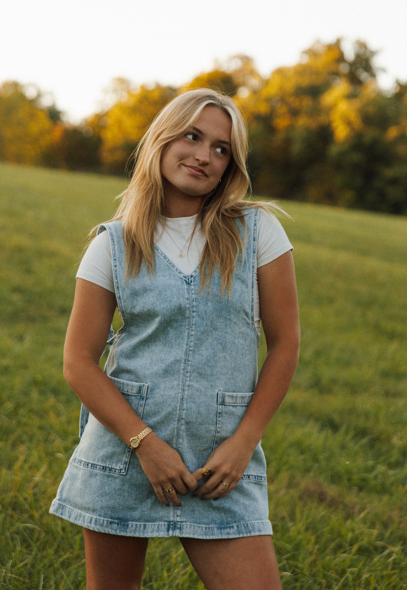 A young woman with blonde hair standing outdoors in a grassy field with trees in the background during fall, wearing a white t-shirt and denim dress, smiling softly.