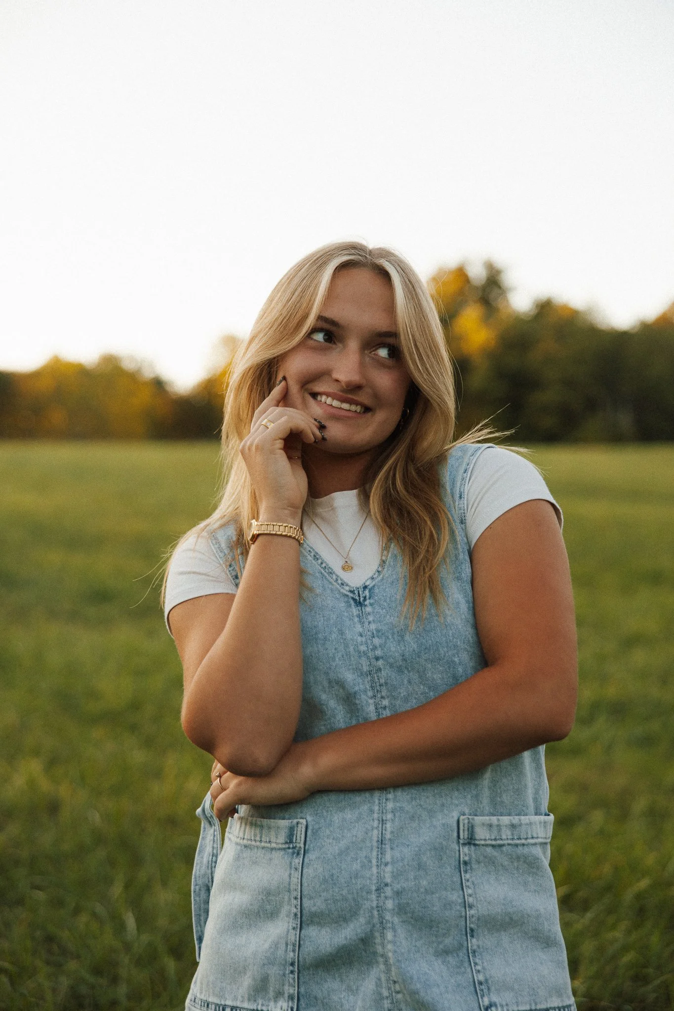A young woman with blonde hair, wearing a white t-shirt and denim overalls, smiling outdoors in a grassy field with trees in the background.