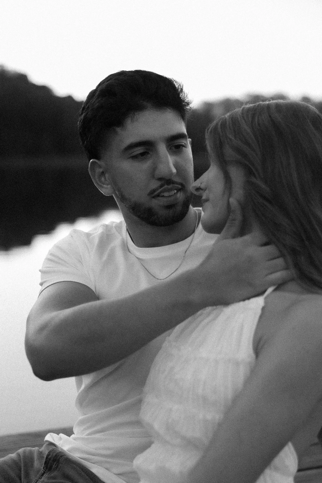 A black and white photo of a young man gently holding a young woman with long hair, by a body of water during sunset or twilight.