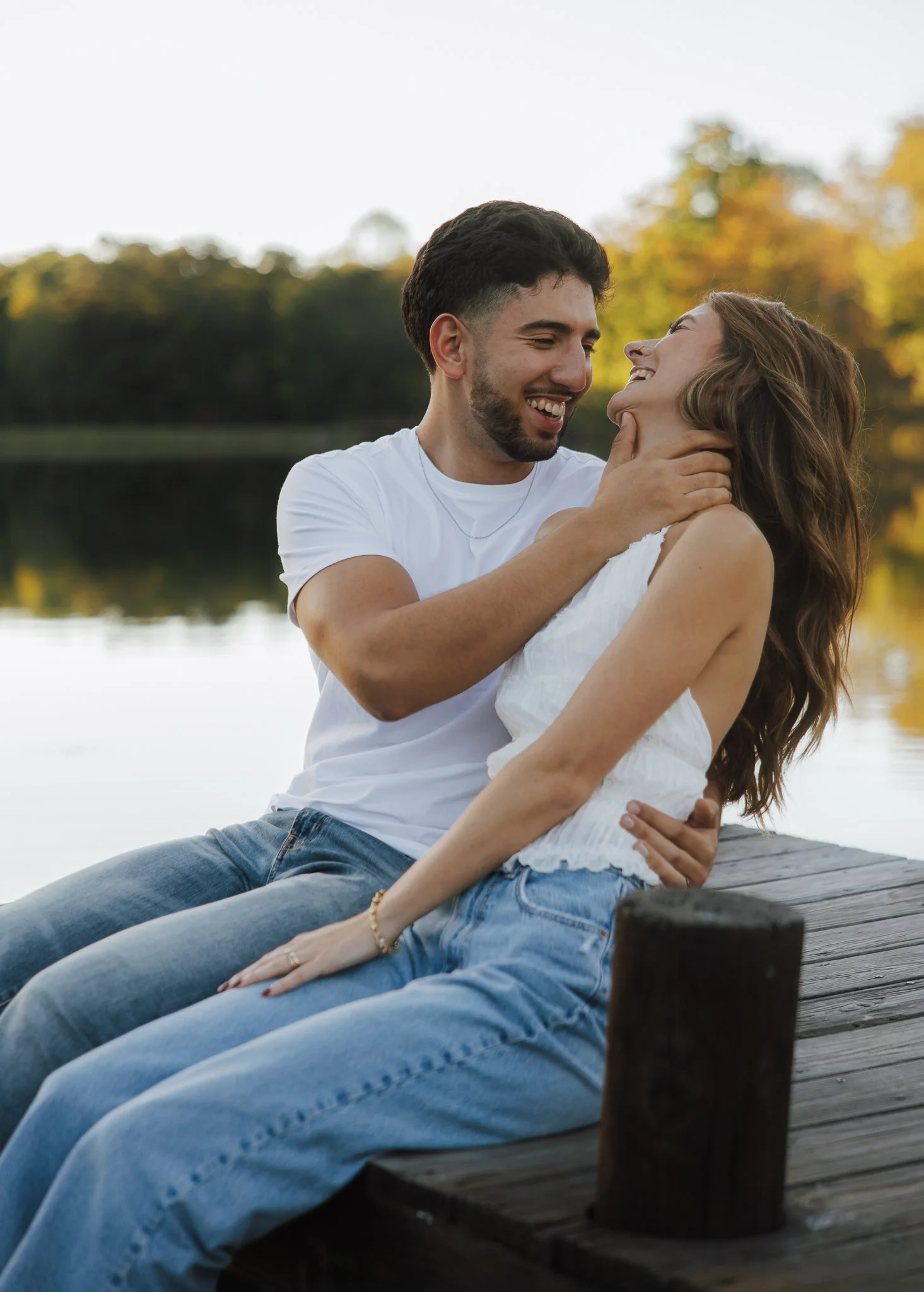 A young couple sitting close together on a wooden dock near a lake, smiling and embracing, with trees showing autumn colors in the background.