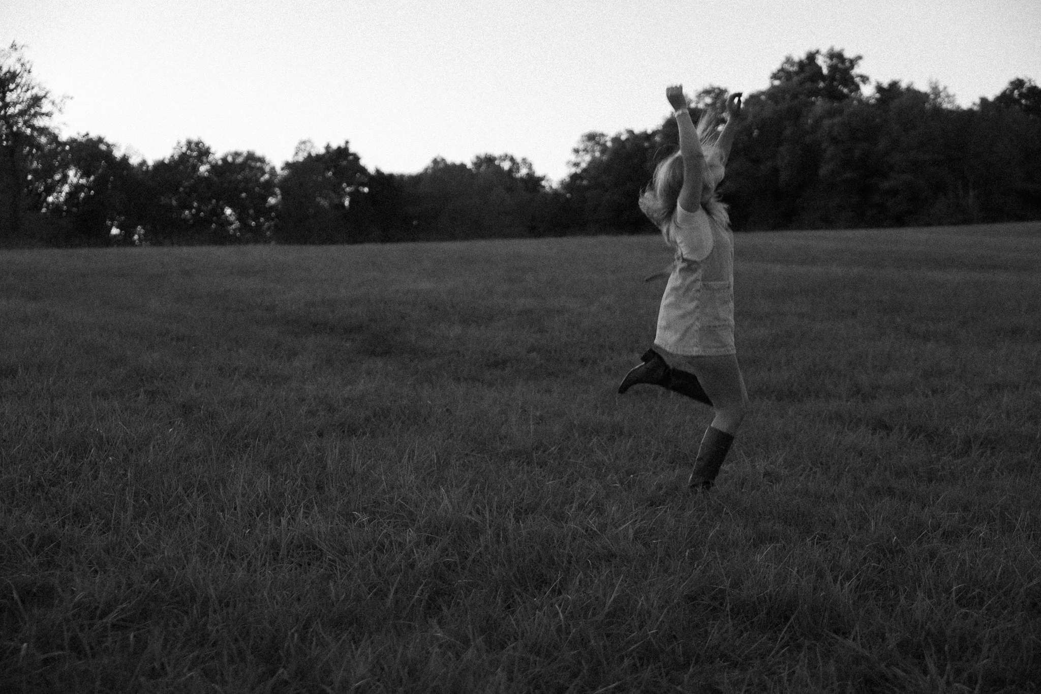 A young girl in a dress and rain boots jumping joyfully in a grassy field at dusk or dawn.