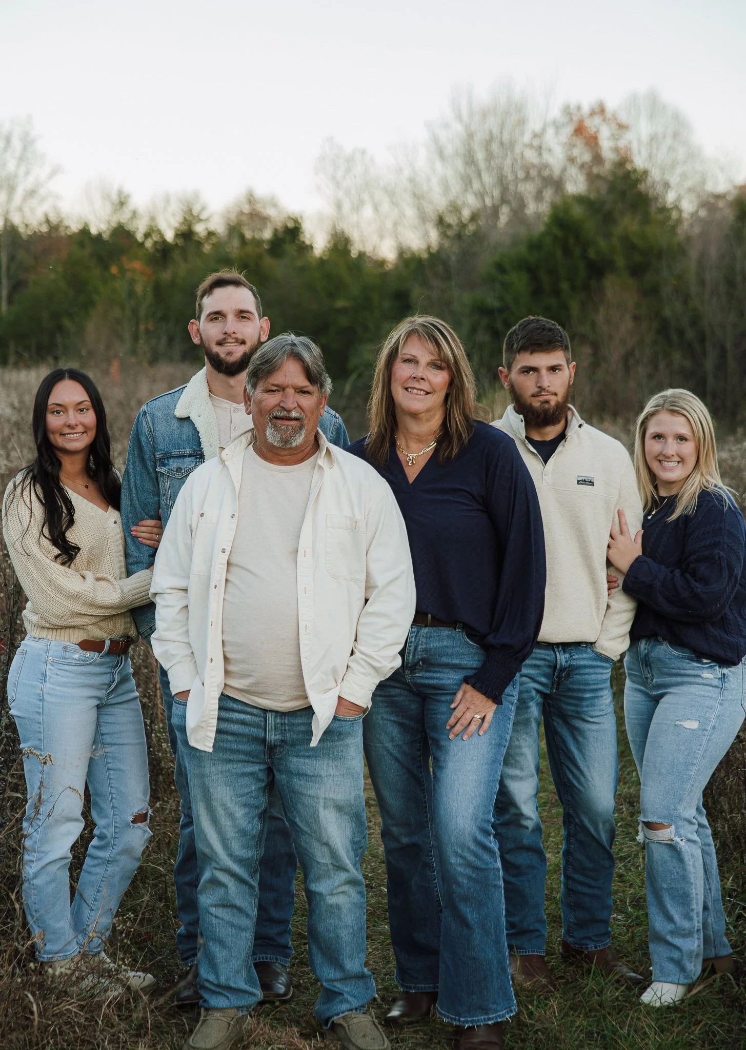 A multi-generational family standing outdoors in a field with trees in the background, smiling at the camera during daytime.