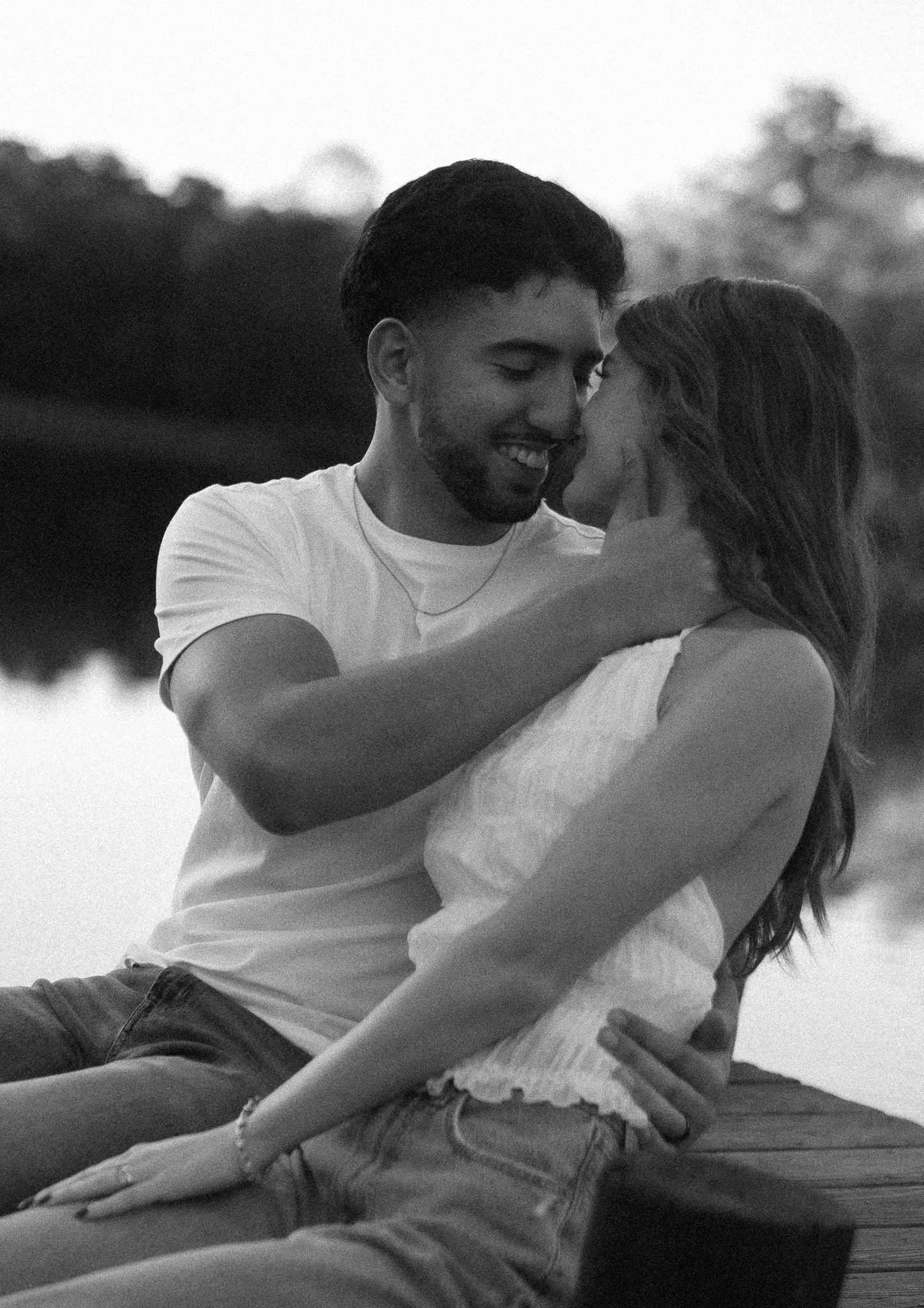 A black and white photo of a couple smiling and embracing near a body of water with trees in the background.