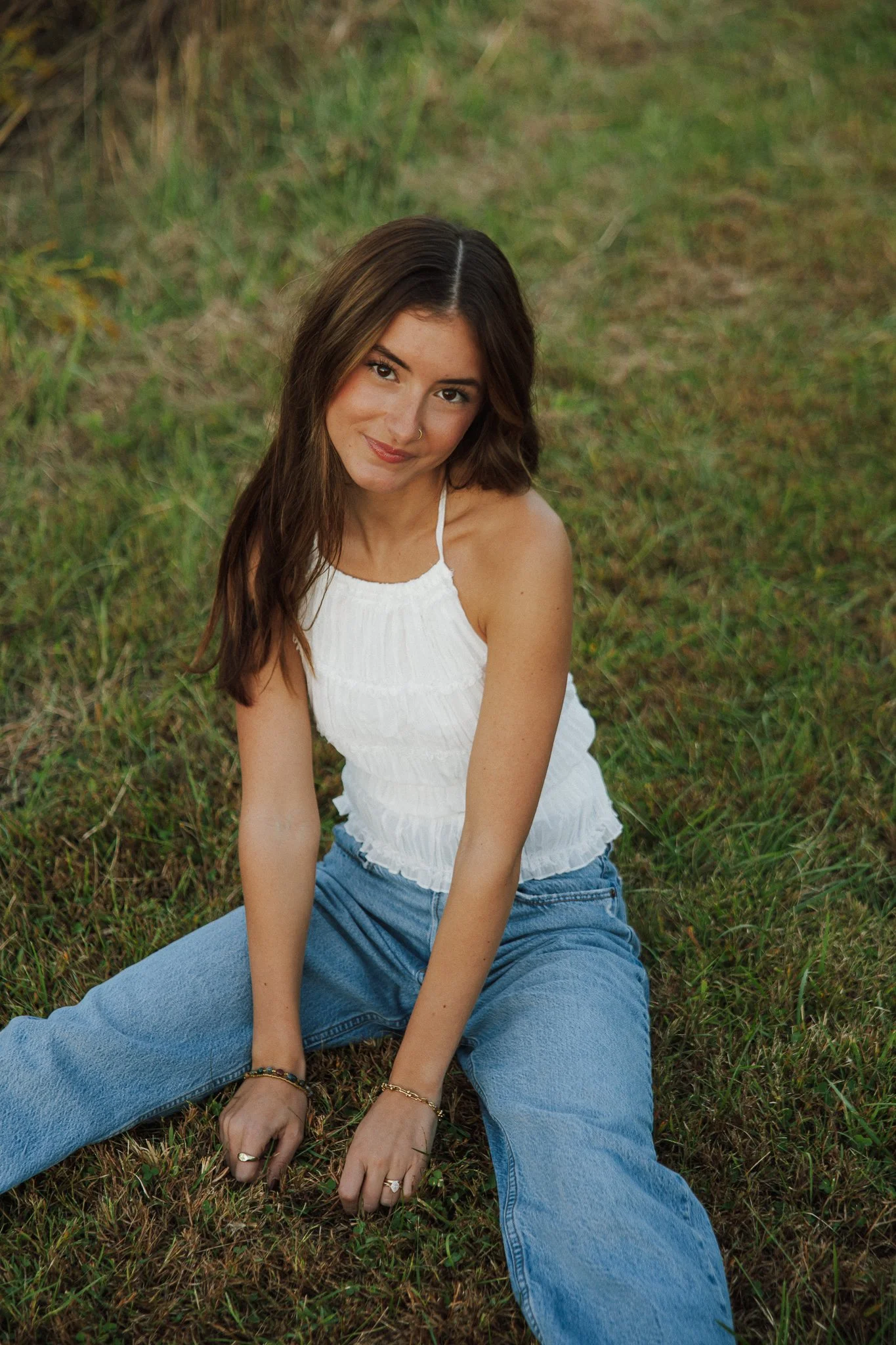 A young woman with brown hair, wearing a white tank top and blue jeans, sitting on grass in a natural outdoor setting, looking at the camera with a slight smile.