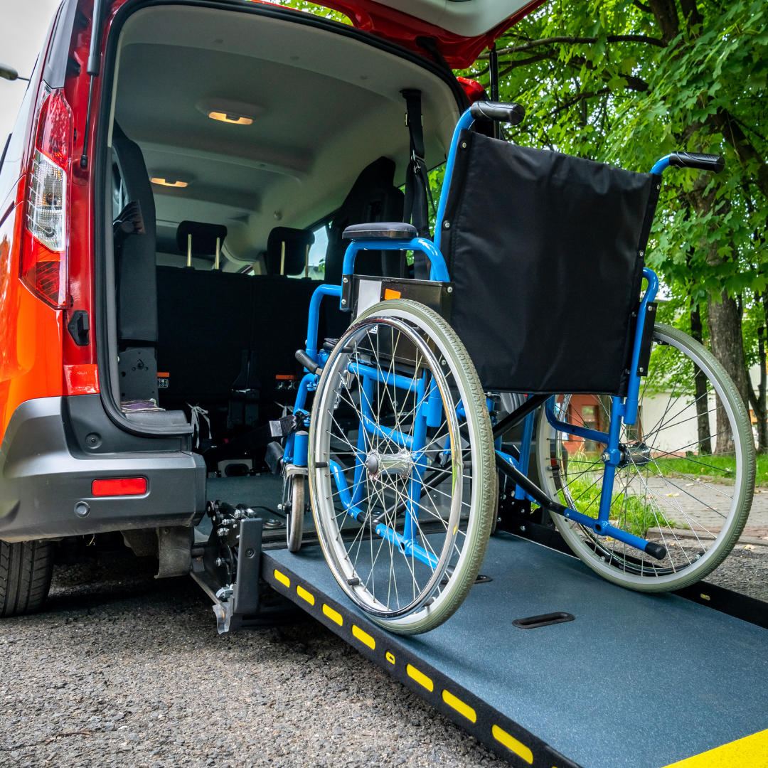 A wheelchair is positioned on a wheelchair accessible ramp lowered from the back of a red vehicle with open doors, outdoors in a green, leafy environment.