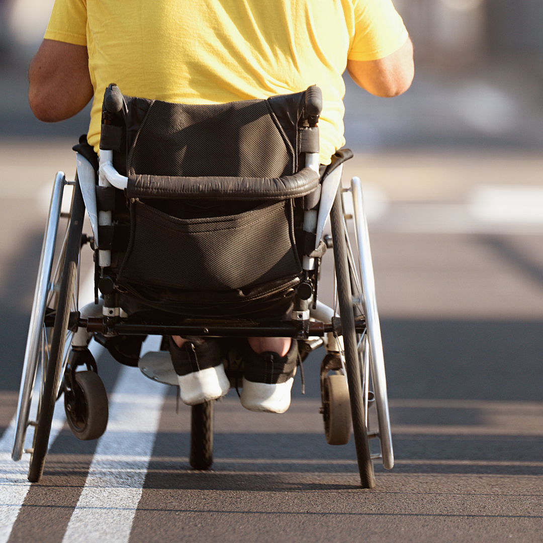 Person in a yellow shirt sitting in a wheelchair on a street, viewed from behind.